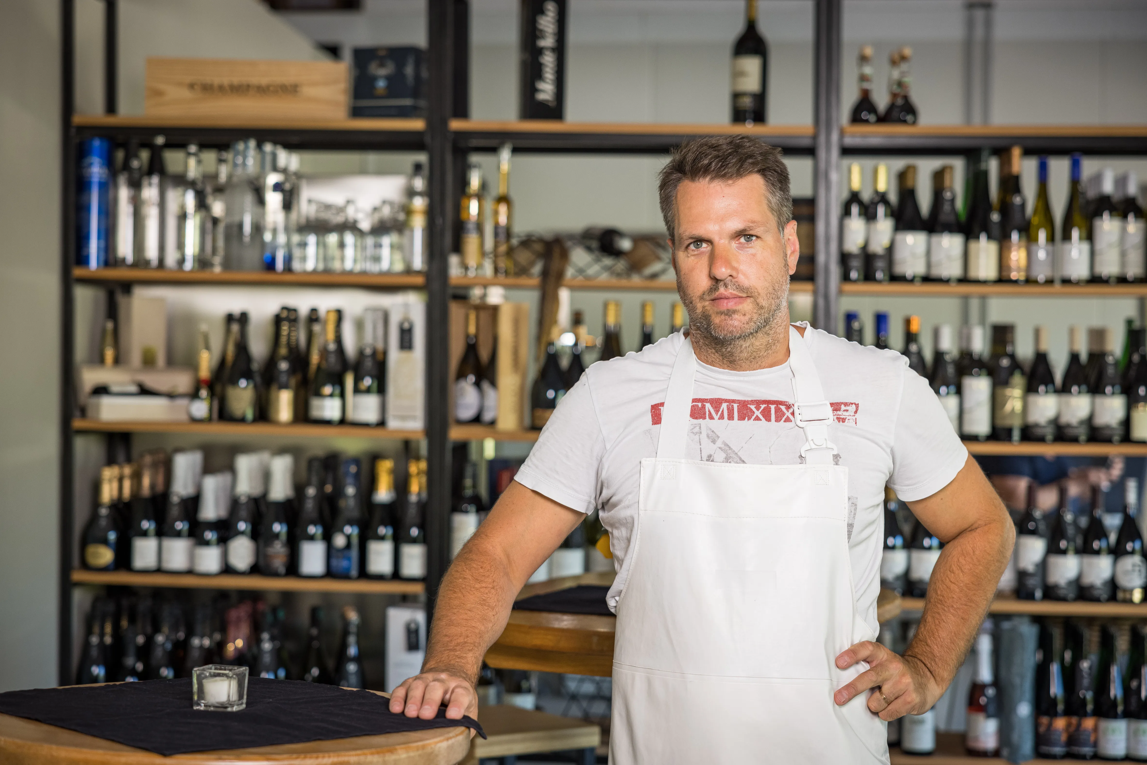 Ein Mann mit weißer Kleidung steht vor einigen Regalen mit verschieden färbigen Flaschen. Daneben ein Stehtisch.