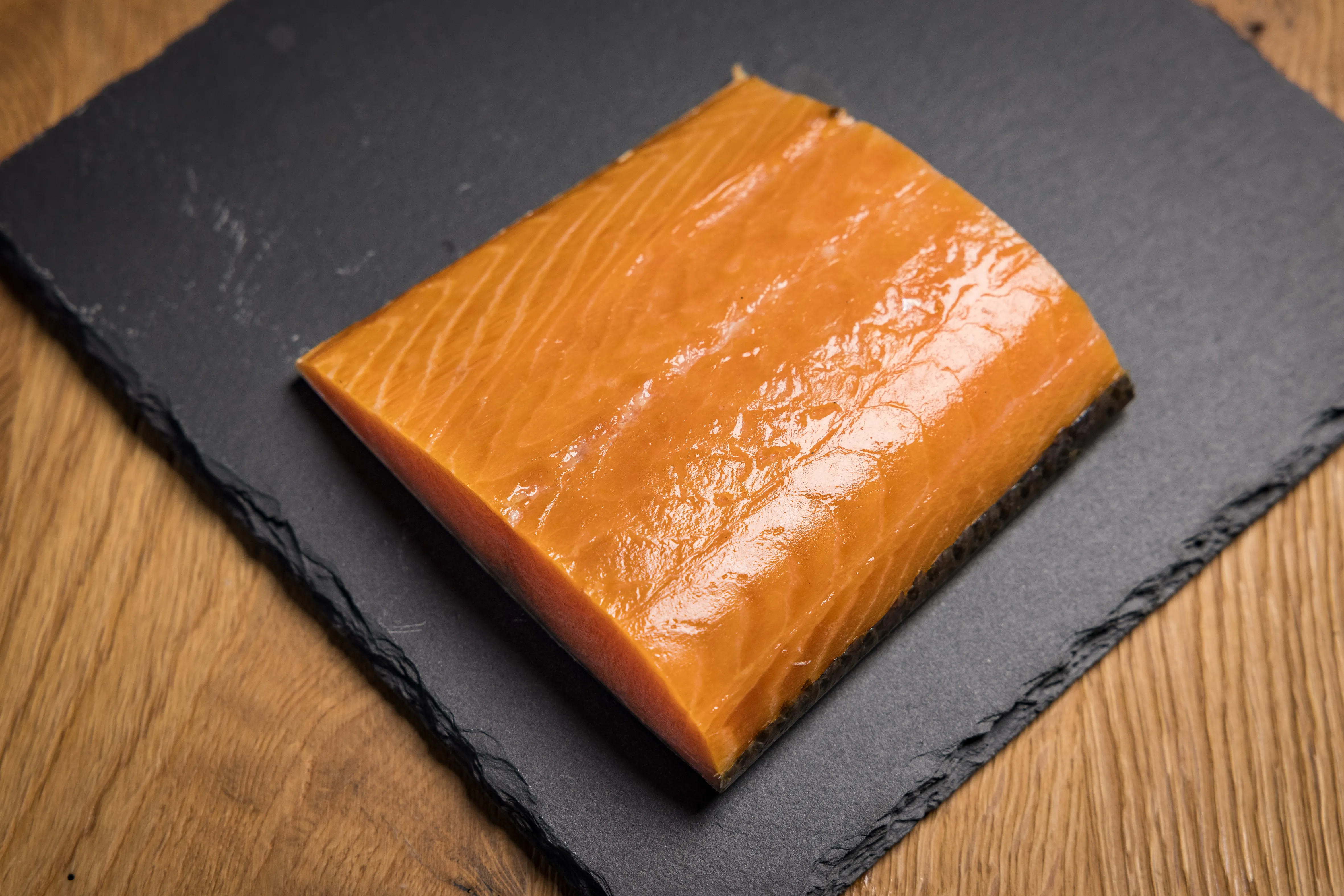 Auf einem braunen Tisch liegt eine schwarze Platte mit einem Stück orangefarbenem Lachs.