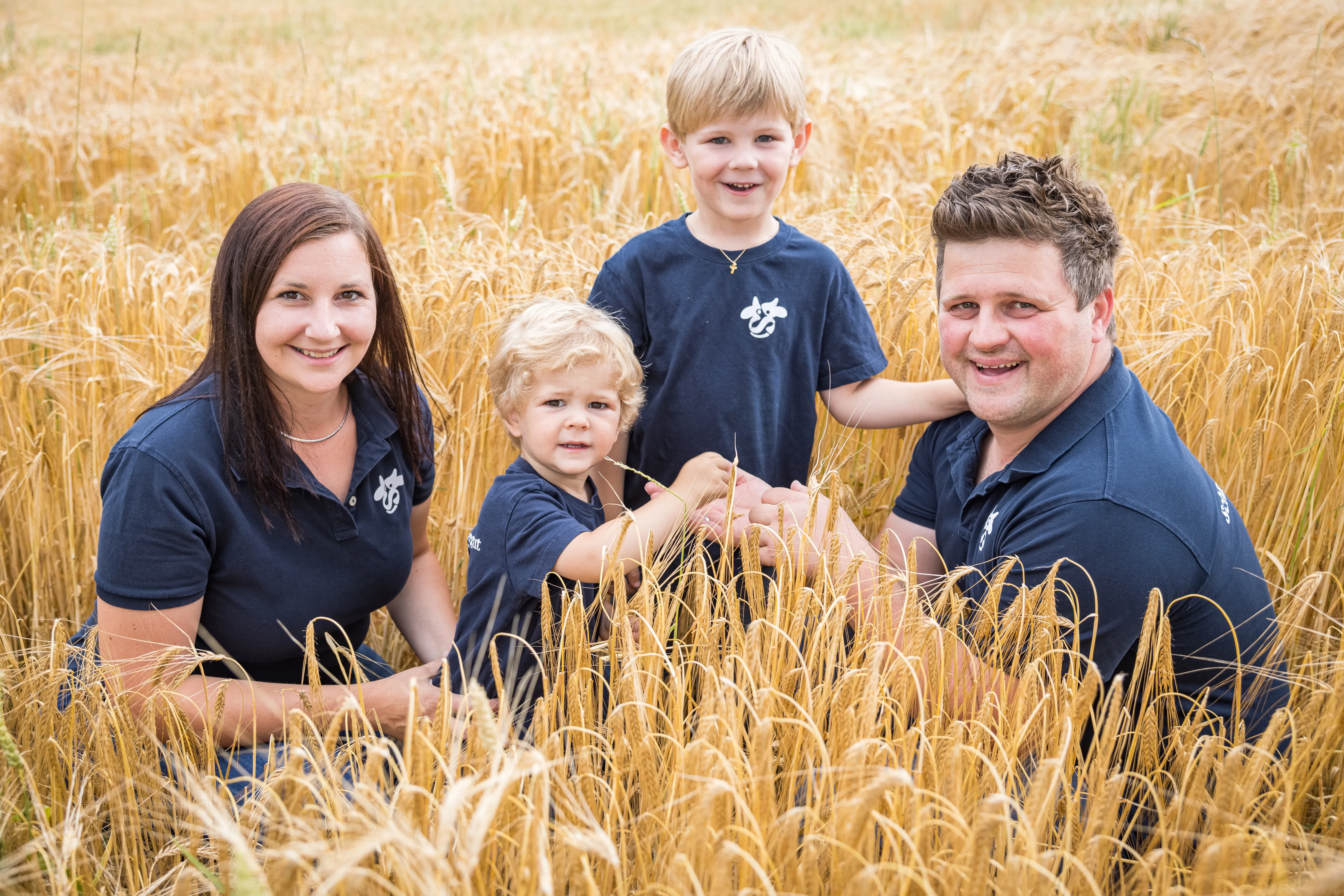 Ein Mann und eine Frau mit zwei Kindern, alle tragen dunkelblaue T-Shirts knieen bzw. stehen in einem goldgelben Feld