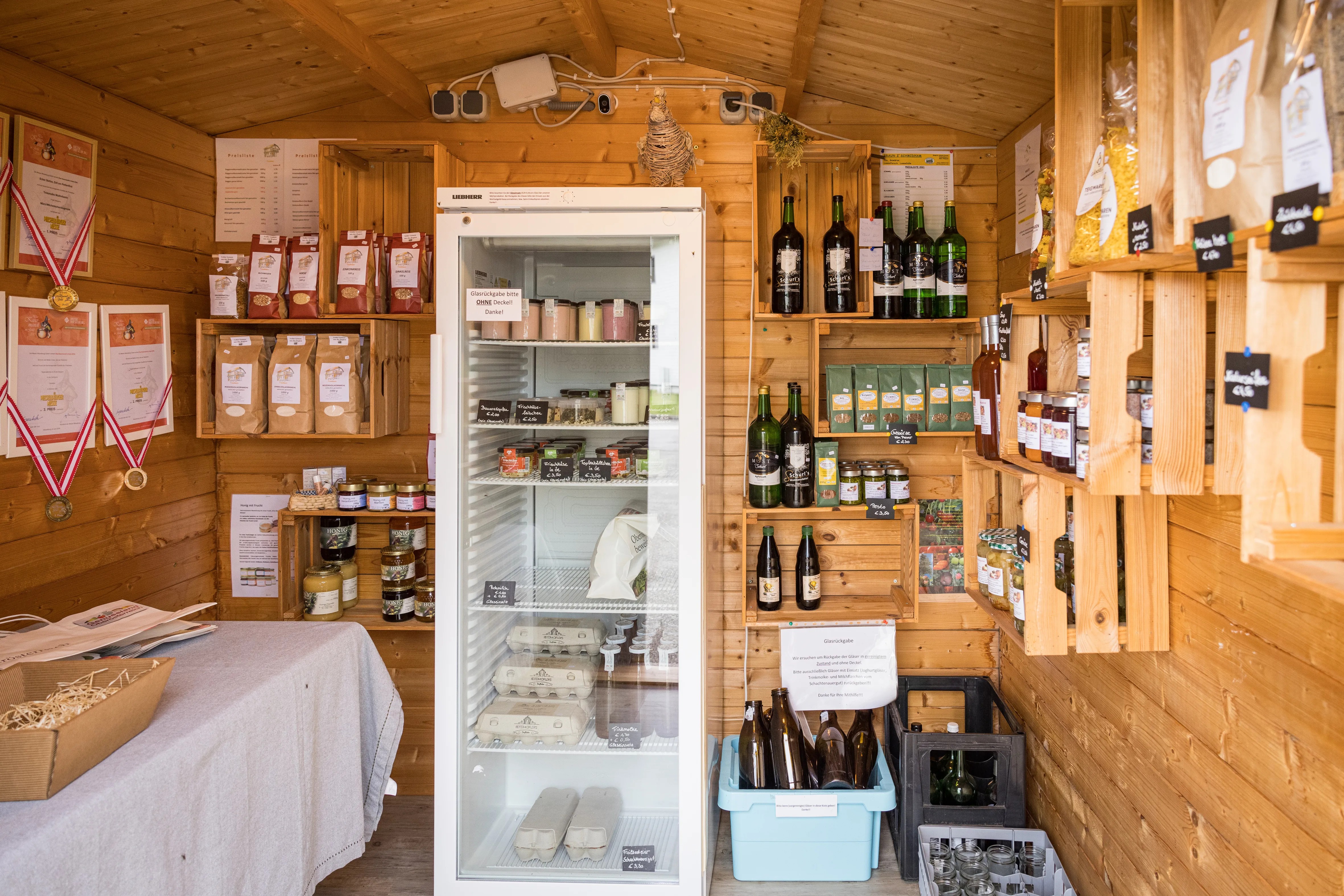 In einer Holzhütte befinden sich mehrere Regale an der Wand mit verschiedenen Produkten wie Flaschen , Gläser und Sackerl. Davor steht ein weißer Kühlschrank mit Glasfenster und ein Tisch mit weißem Tischtuch. An der Wand hängen mehrere Urkunden und Medaillen.