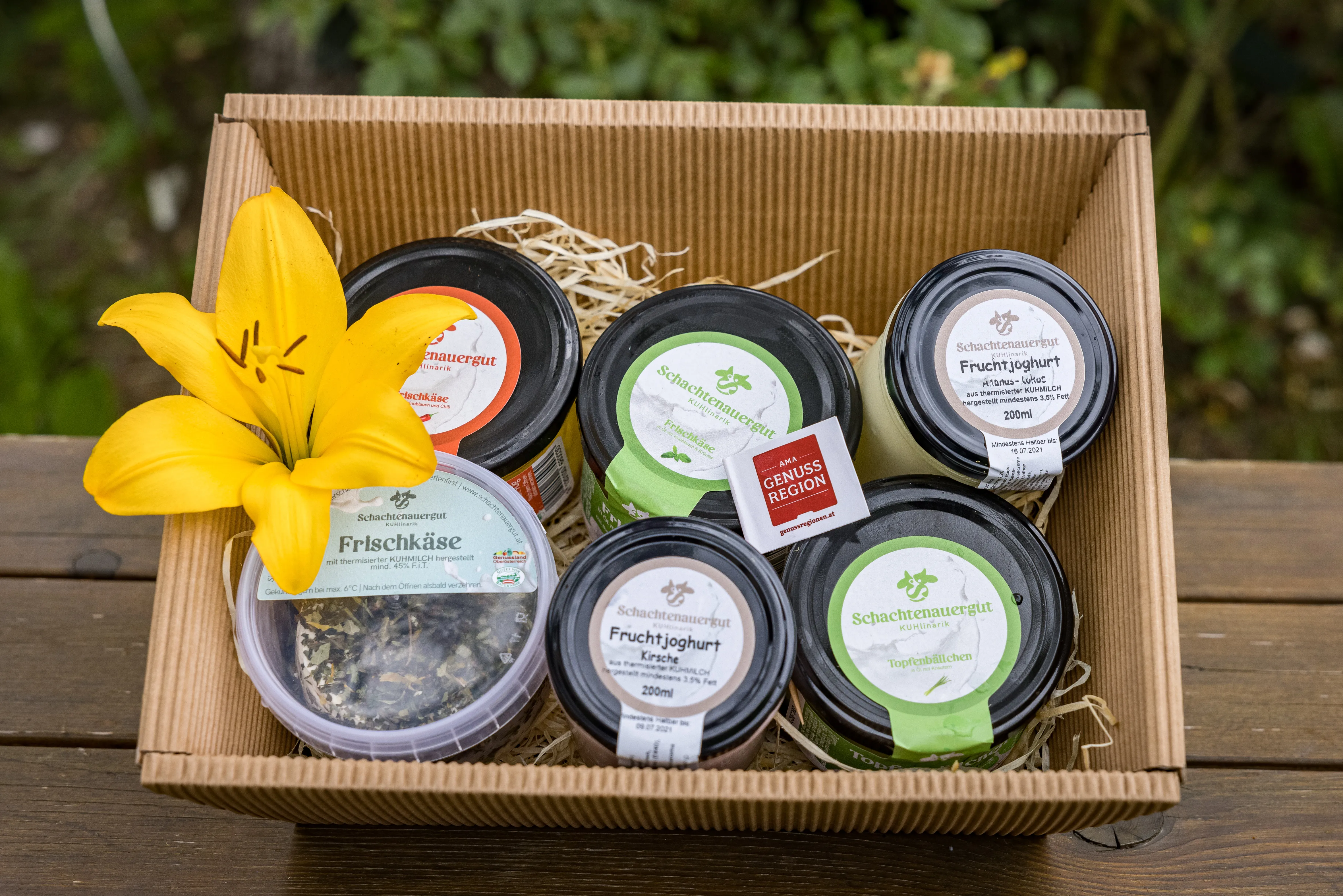 Auf einem Holztisch steht eine braune Kiste mit verschiedenen Gläsern. Dazwischen steckt eine gelbe Lilie. Dahinter grüne Wiese