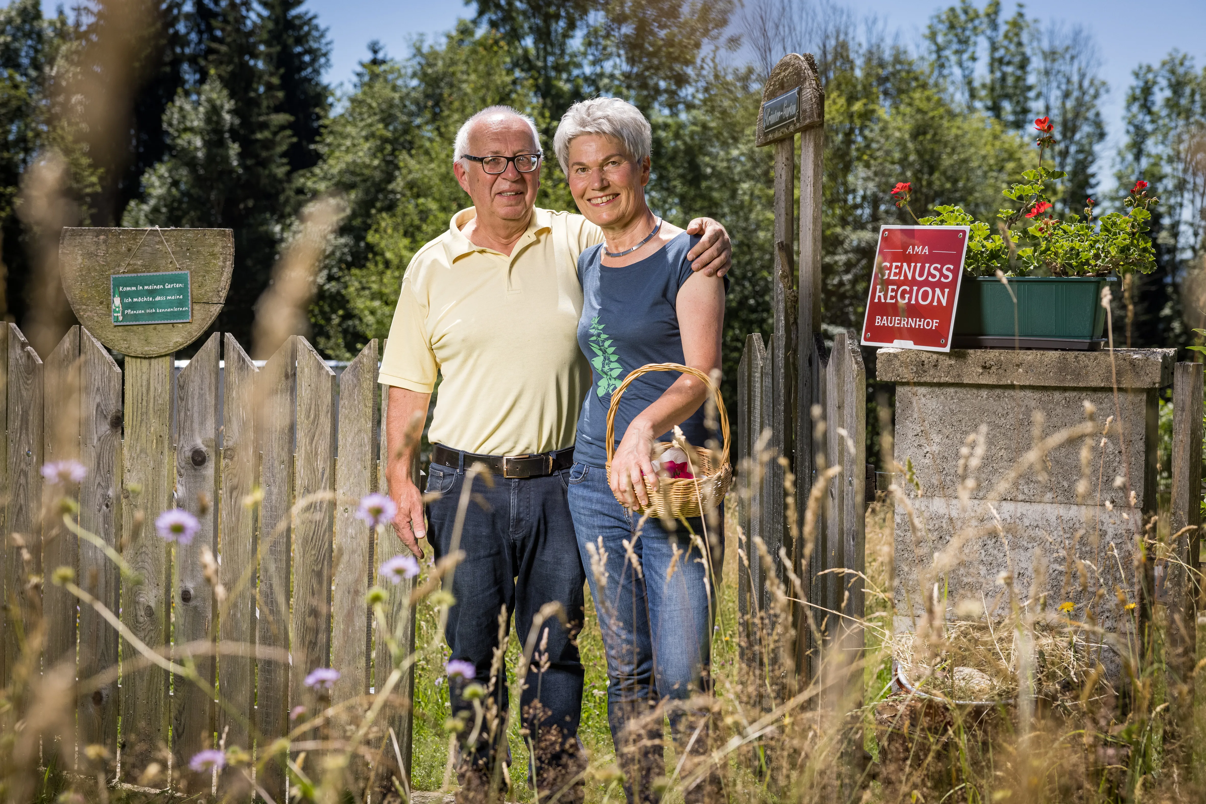 Ein Mann und eine Frau stehen mit einem Korb in der Hand vor einem Holzzaun. Davor blühende Wiese, dahinter grüne Bäume und blauer Himmel. Der Mann hält die Frau an der Schulter.