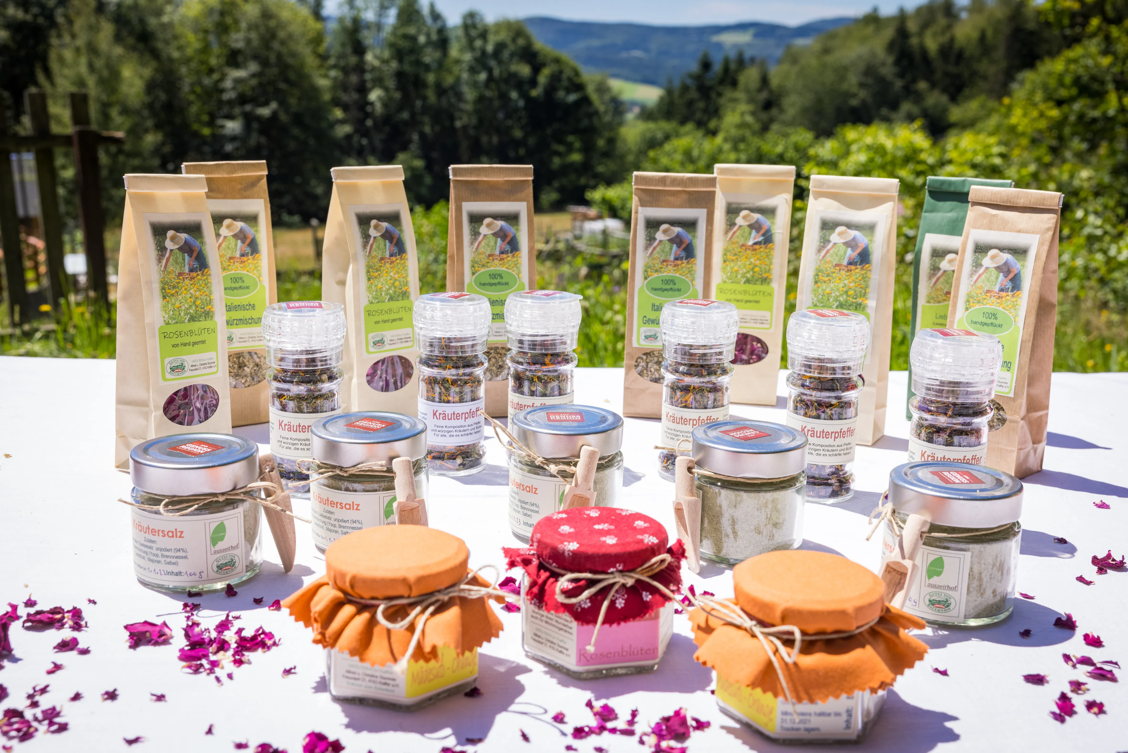 Auf einem weißen Tisch stehen verschieden färbige Gläser und Sackerl mit Kräuter und Kräuterprodukte. Dahinter grüne Wiese und grüner Wald.