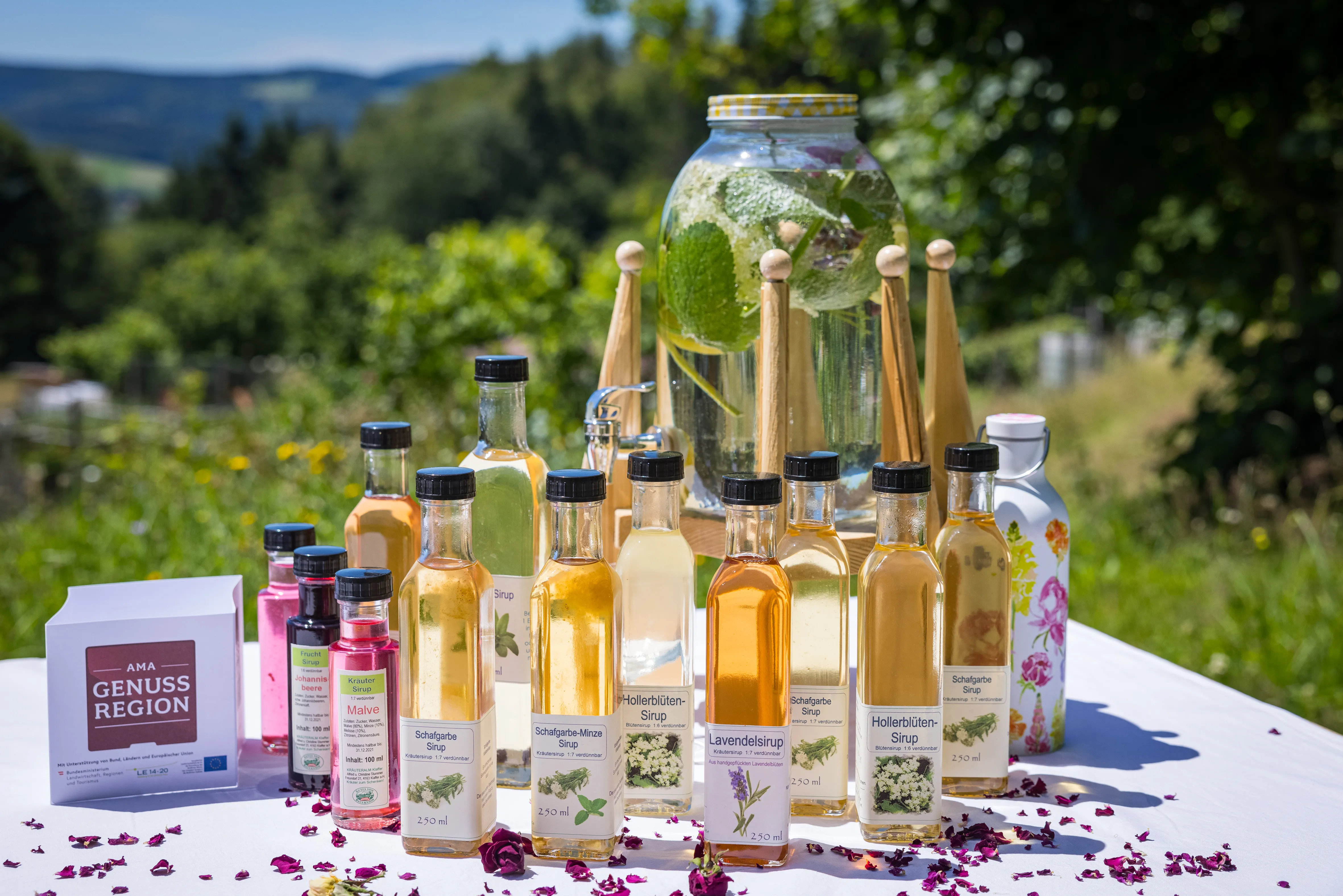 Auf einem weißen Tisch stehen verschieden große Flaschen mit verschieden färbigem Inhalt, daneben ein Kartonwürfel mit der Aufschrift AMA Genuss Region, dazwischen Blüteblätter und Kräuter. Dahinter grüne Wiese und Wald.