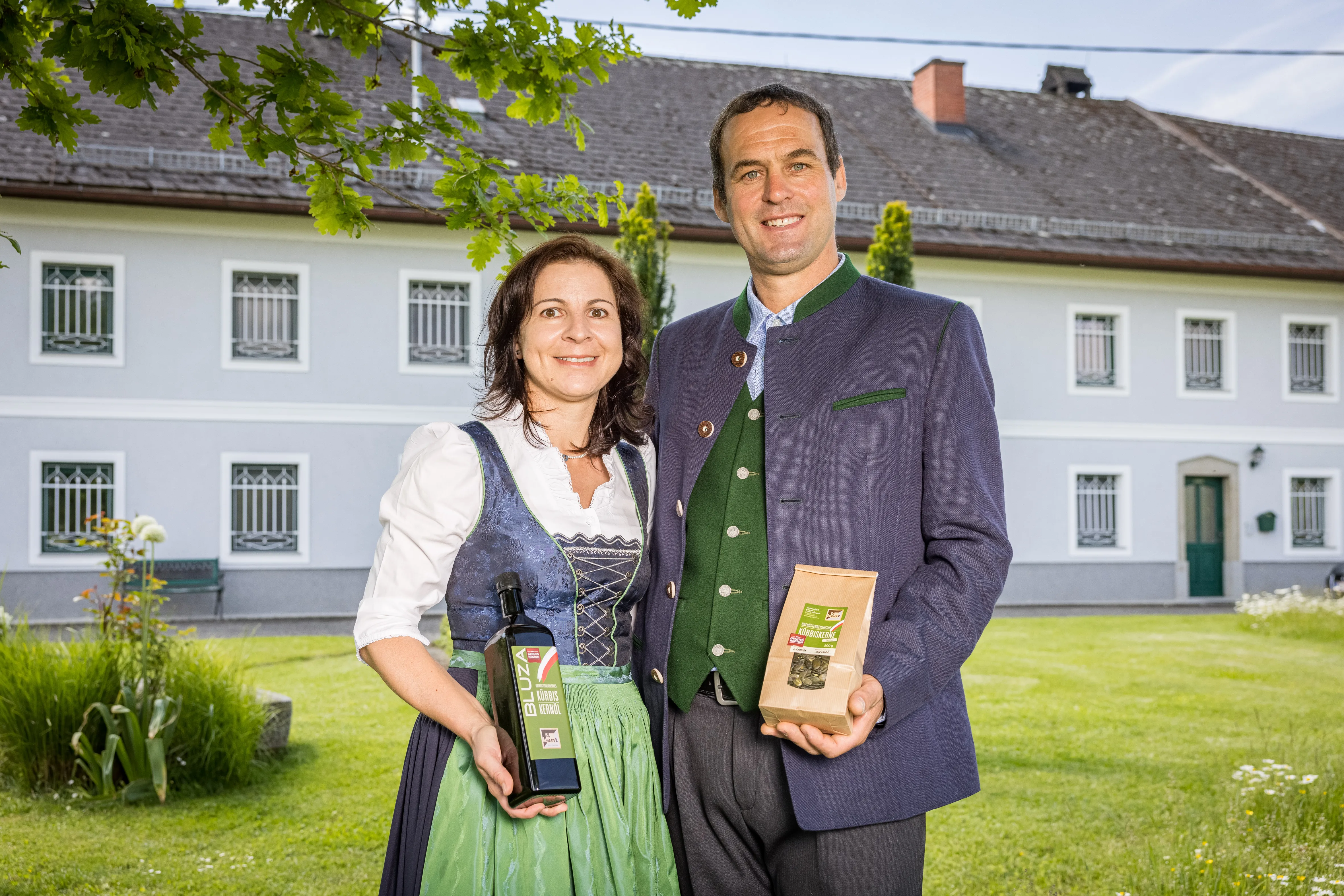 Eine Frau im blau/grünen Dirndl mit weißer Bluse und ein Mann im grau/grünen Anzug stehen vor einem großen Bauernhof, hellblau/weiße Fassade auf einer grünen Wiese. Sie halten eine Flasche und ein Sackerl Kerne in den Händen.