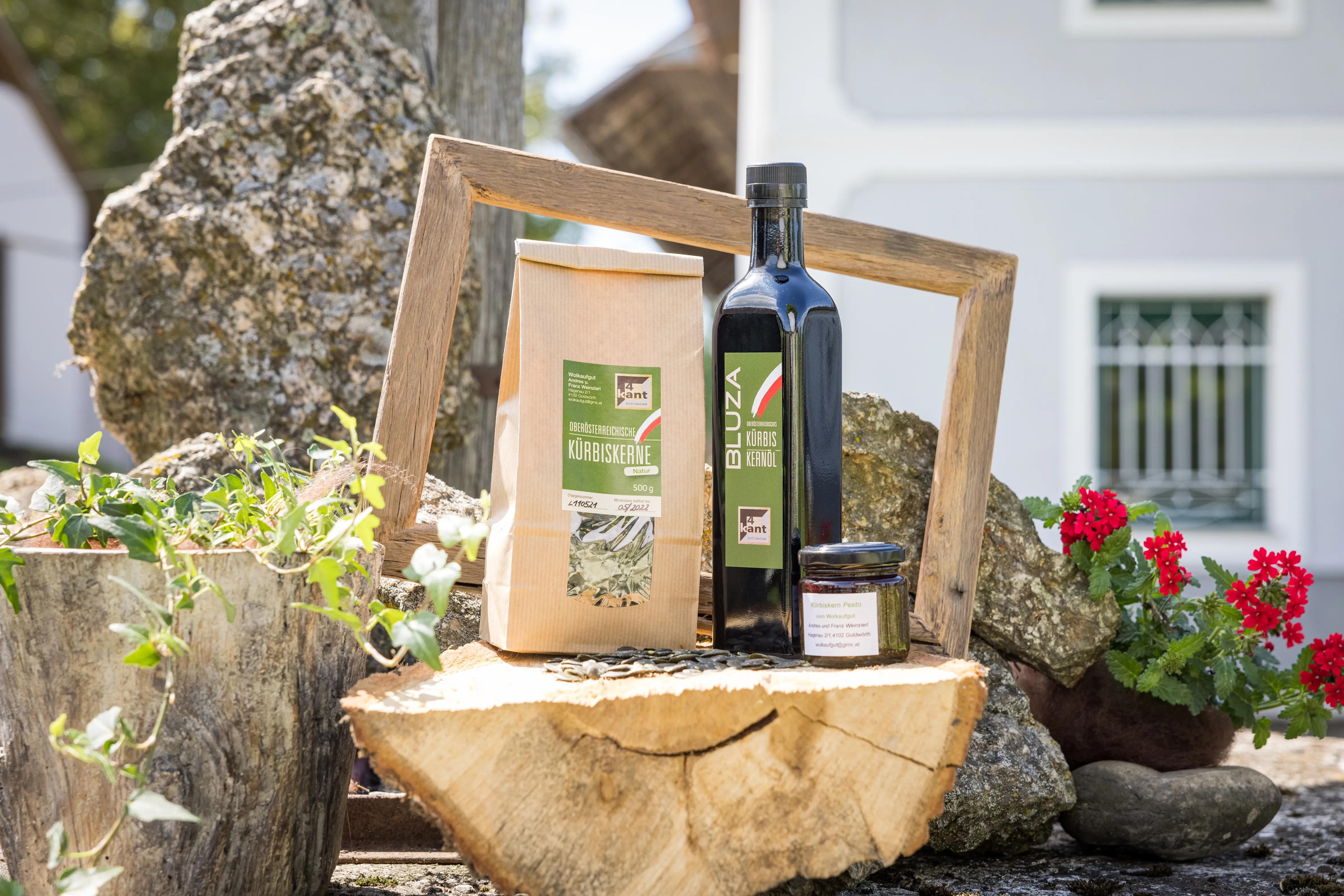 Auf einem Holzstumpf stehen eine grüne Flasche, ein grünes Glas und ein Papiersackerl. Dahinter ein Stein und eine hellblaue Fassade eines Bauernhofes. Daneben rote und grüne Pflanzen.