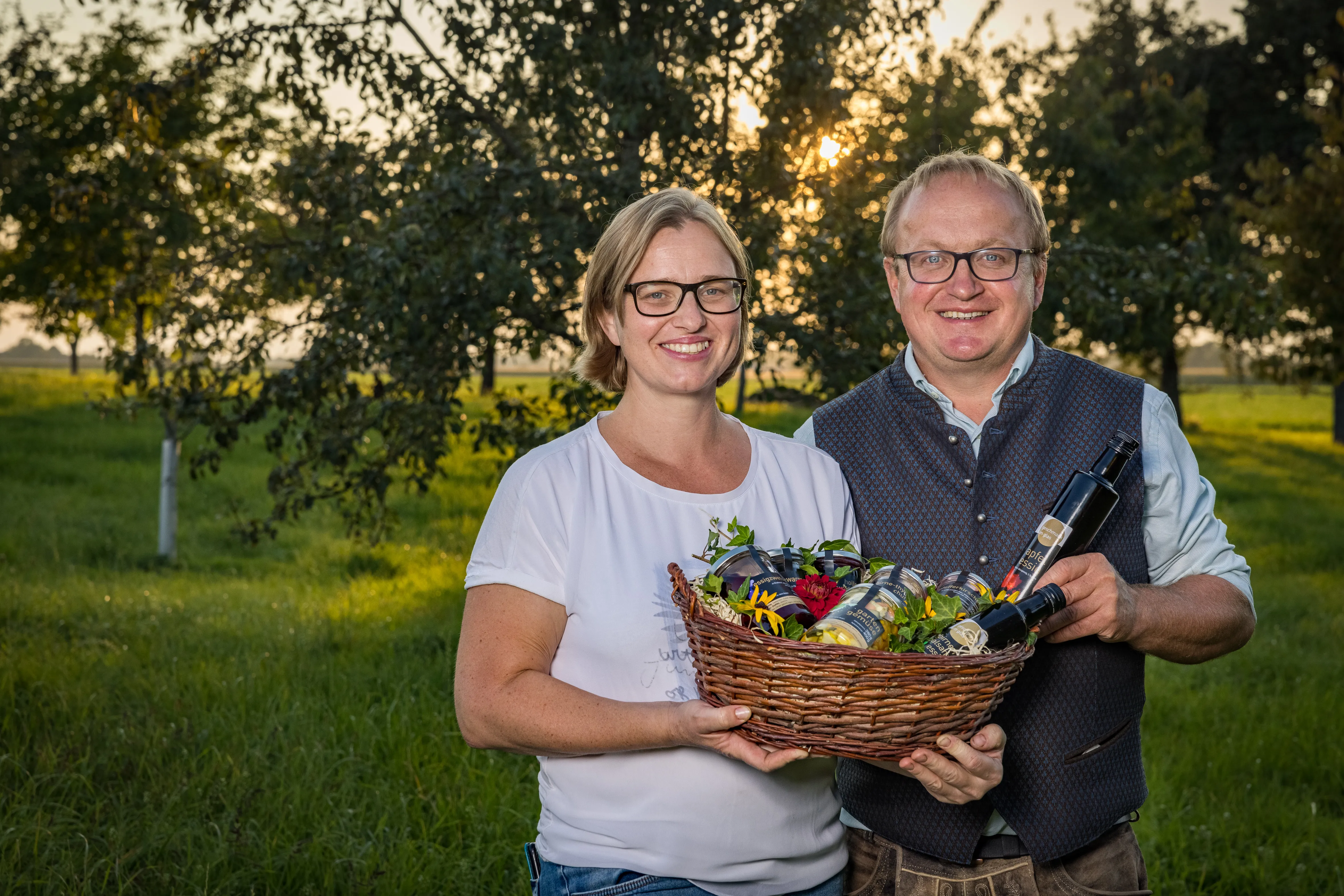 Ein Mann und eine Frau stehen auf einer grünen Wiese, dahinter Bäume, das Sonnenlicht scheint zwischen den Bäumen durch; die beiden halten einen Korb mit Produkten in den Händen. Beide sind Brillenträger.