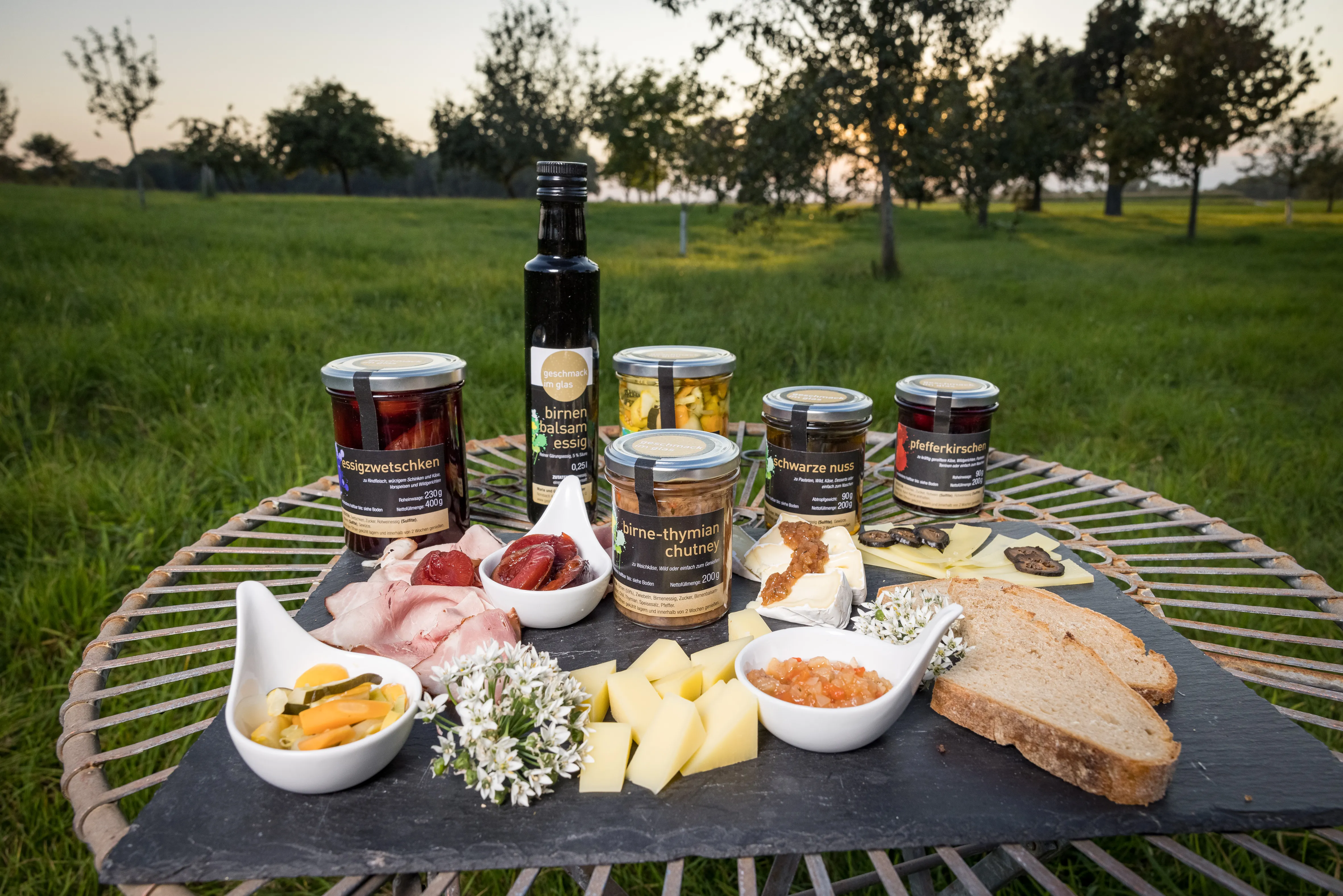 Auf einer grünen Wiese steht ein tisch mit einer schwarzen Platte. Darauf stehen Gläser, eine Flasche, verschiedene Schalen mit Eingelegtem, dazwischen aufgeschnittener Käse und Brotscheiben und Schinken. Im Hintergrund grüne Bäume und Wiese.