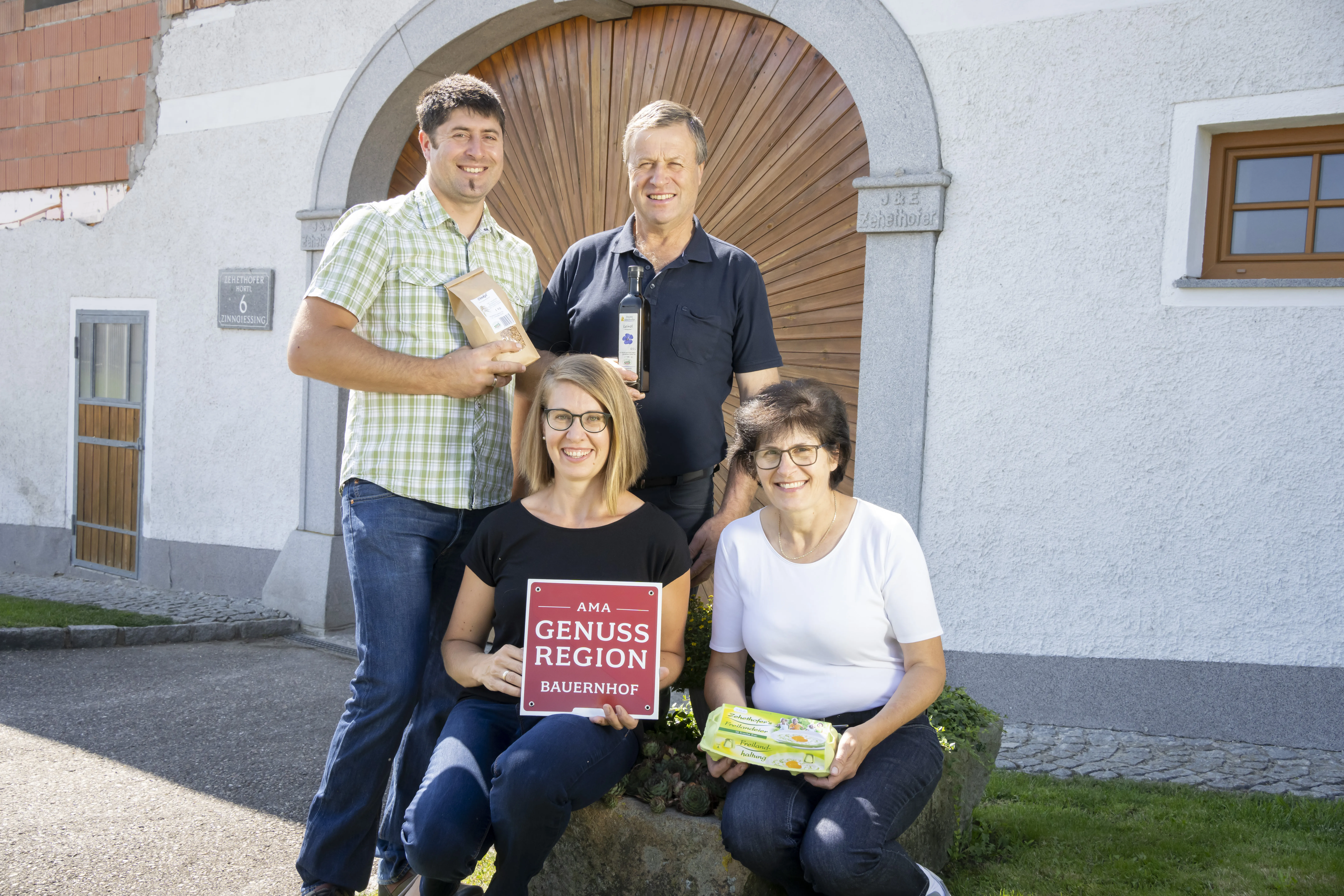 Vor einem weißen Haus mit braunem Hoftor stehen ein älterer Mann mit schwarzem Hemd und ein jüngerer Mann mit einem grün/weiß gestreiften Hemd, davor knieen zwei Frauen, eine mit einem schwarzen T-Shirt und eine ältere mit einem weißen T-Shirt. Daneben Asphalt und grüne Wiese