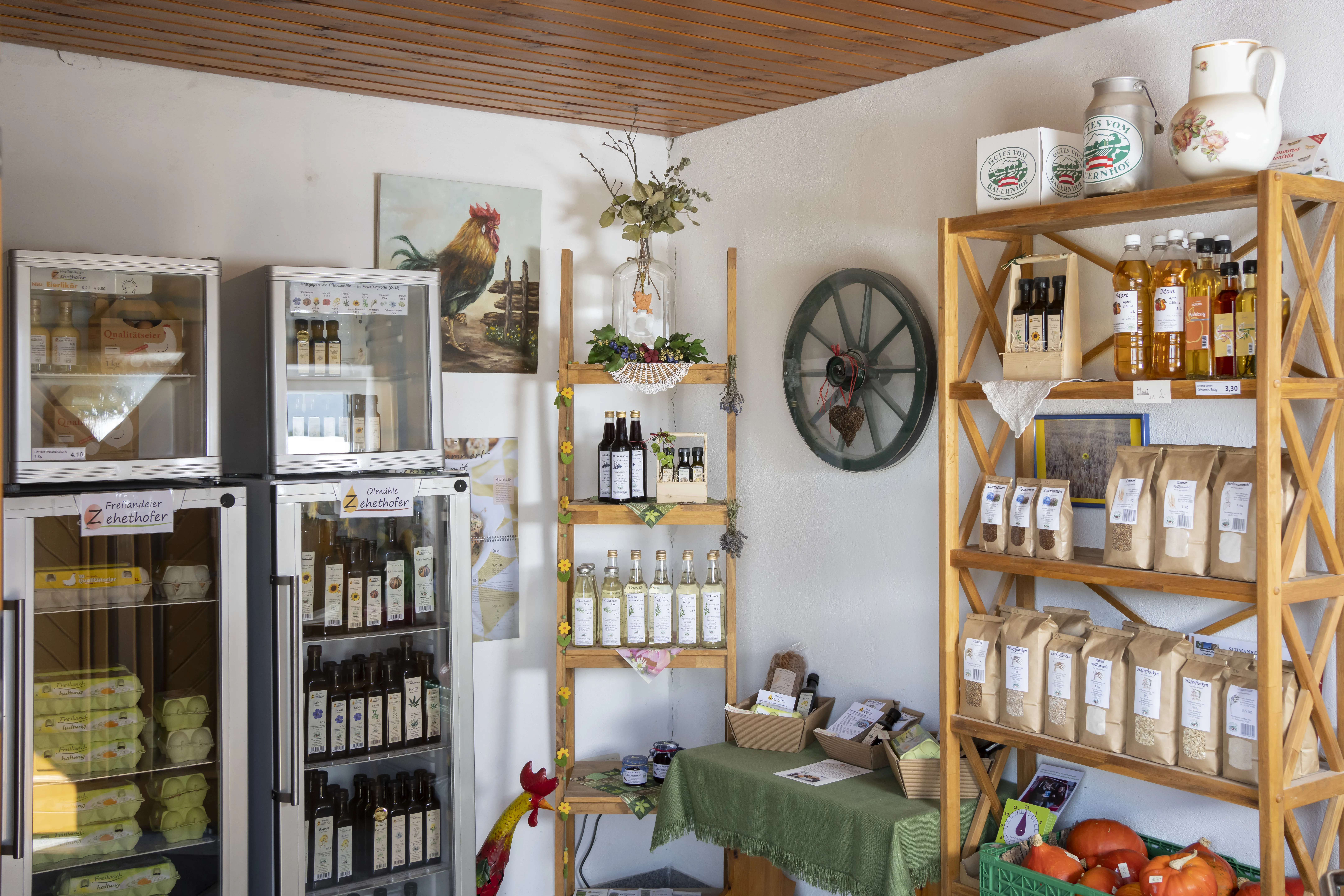 An einer weißen Wand stehen weiße Kühlschränke und braune Regale mit verschiedenen Produkten darauf (Flaschen, Gläser, Sackerl. Ein Bild mit einem Hahn hängt an der Wand, ein braunes Karrenrad und ein kleiner Tisch mit grüner Tischdecke