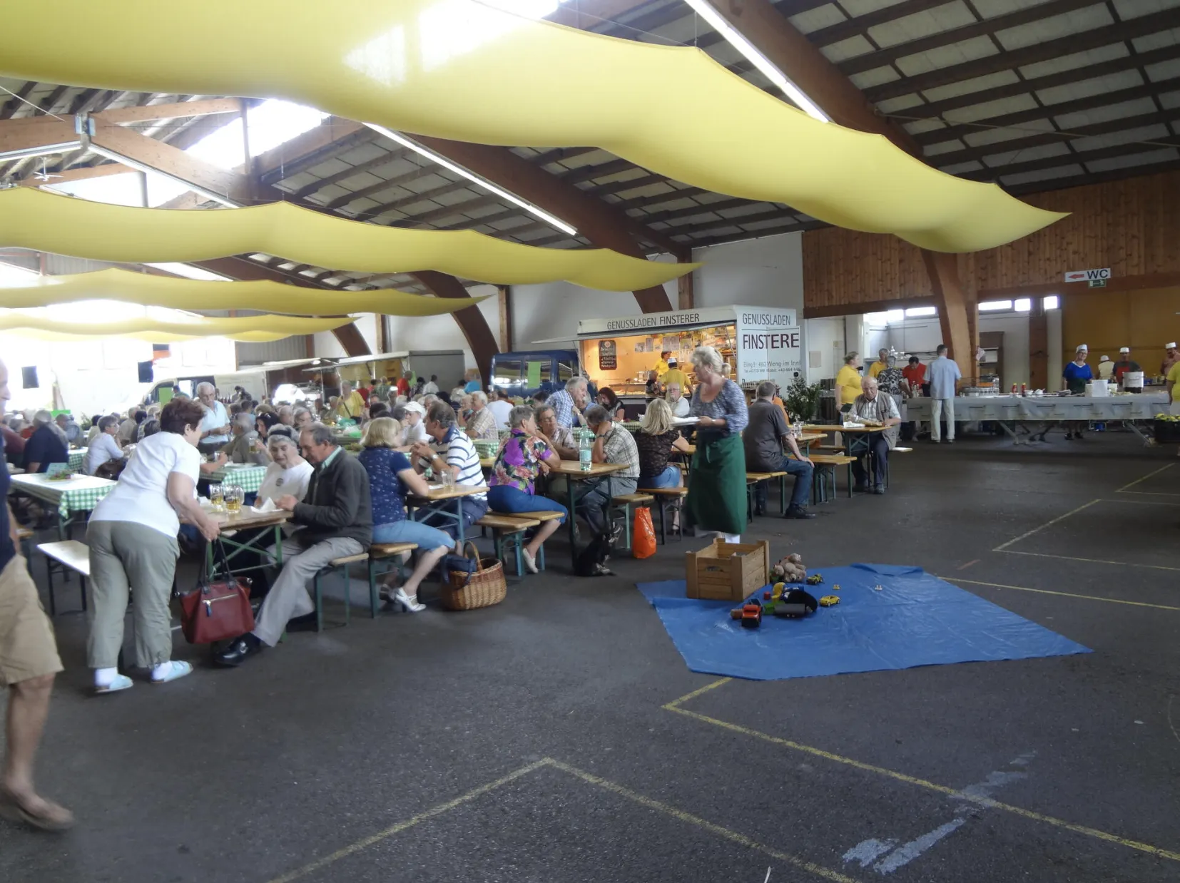 Ein großer Raum mit velen Tischen, an denen zahlreiche Menschen sitzen und essen. An den Decken hängen gelbe Luftballons. In einer Ecke des Raums befindet sich ein Verkaufsstand mit Hinweisschildern darauf.