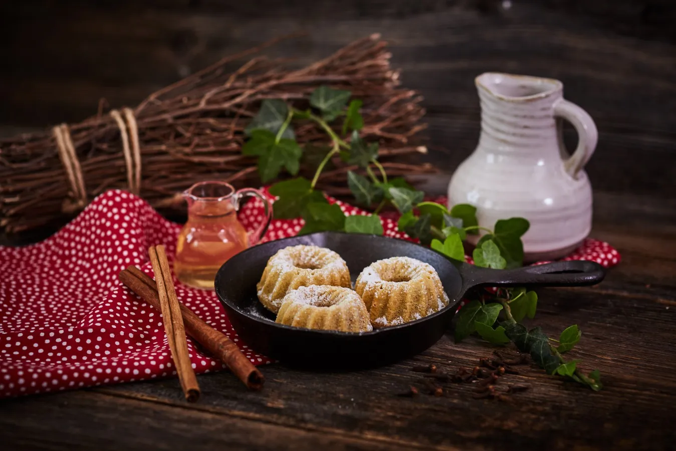 Mostpudding - In einer schwarzen Pfanne befinden sich drei goldbraune Stück Mostpudding. Die Pfanne steht auf einem braunen Holztisch und ist mit einem roten Tischtuch mit weißen Punkten unterlegt. Links neben der Pfanne liegen zwei Zimtstangen. Links hinter der Pfanne befindet sich ein kleiner Glaskrug, der mit Most gefüllt ist. Rechts hinter der Pfanne ist ein weißer Tonkrug zu sehen. Grünes Efeu und ein braunes Bündel Holz dienen der Zierde.