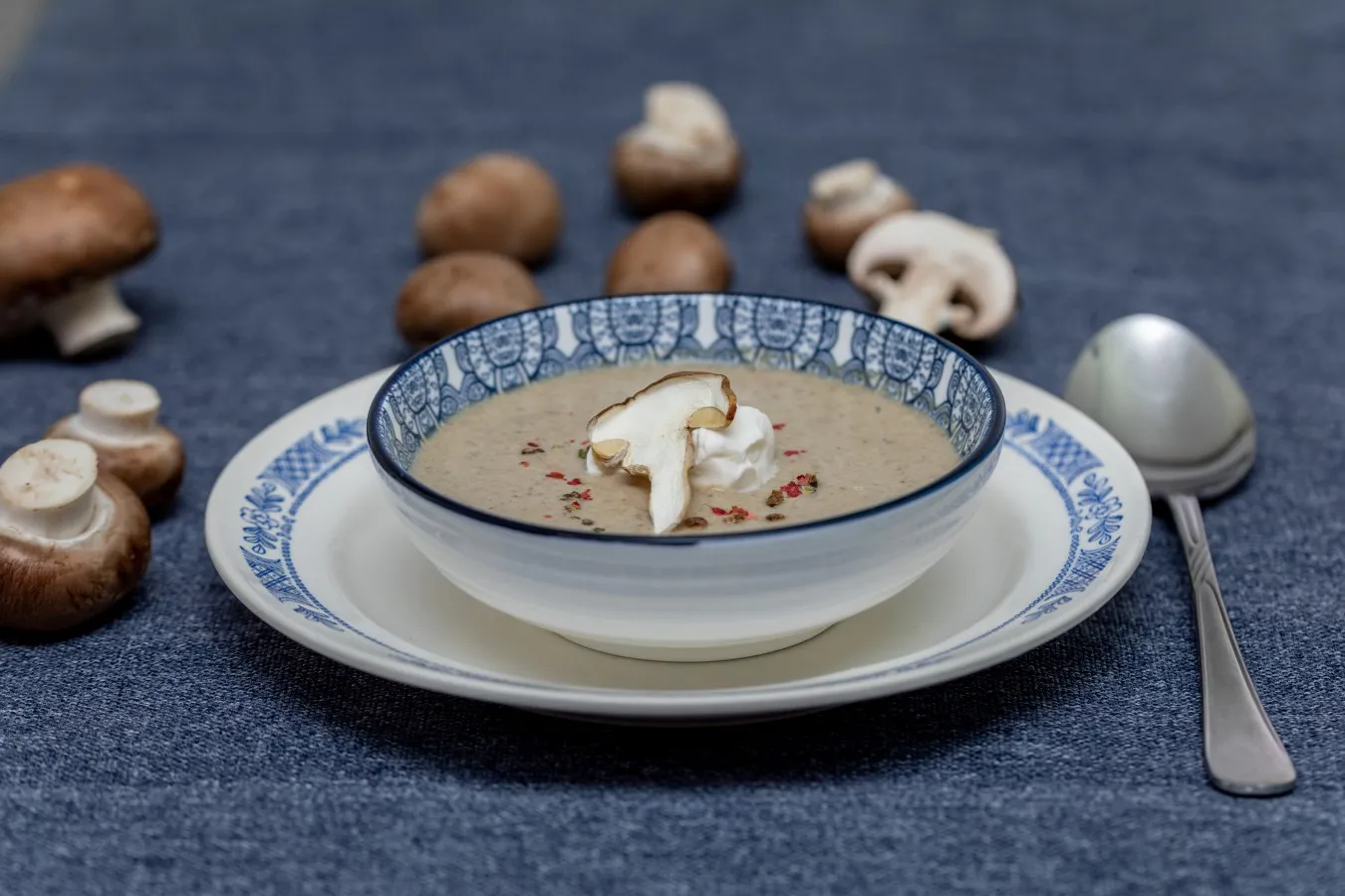 Pilzcremesuppe - Auf blauem Untergrund steht ein weißer Teller mit blauer Verziehrung. Auf dem Teller befindet sich eine Suppenschüssel ebenfalls mit blauer Verziehrung. In der Suppenschüssel ist die hellbraune Pilzcremesuppe angerichtet. Die Suppe ist in der Mitte mit einem Tupfen Schlagobers einem Stück Champignon und bunten Pfefferkörnern garniert. Auf der linken Seite und hinter dem Teller liegen einige braune Chamoignons verteilt. Rechts neben dem Teller liegt ein silbener Suppenlöffel. 