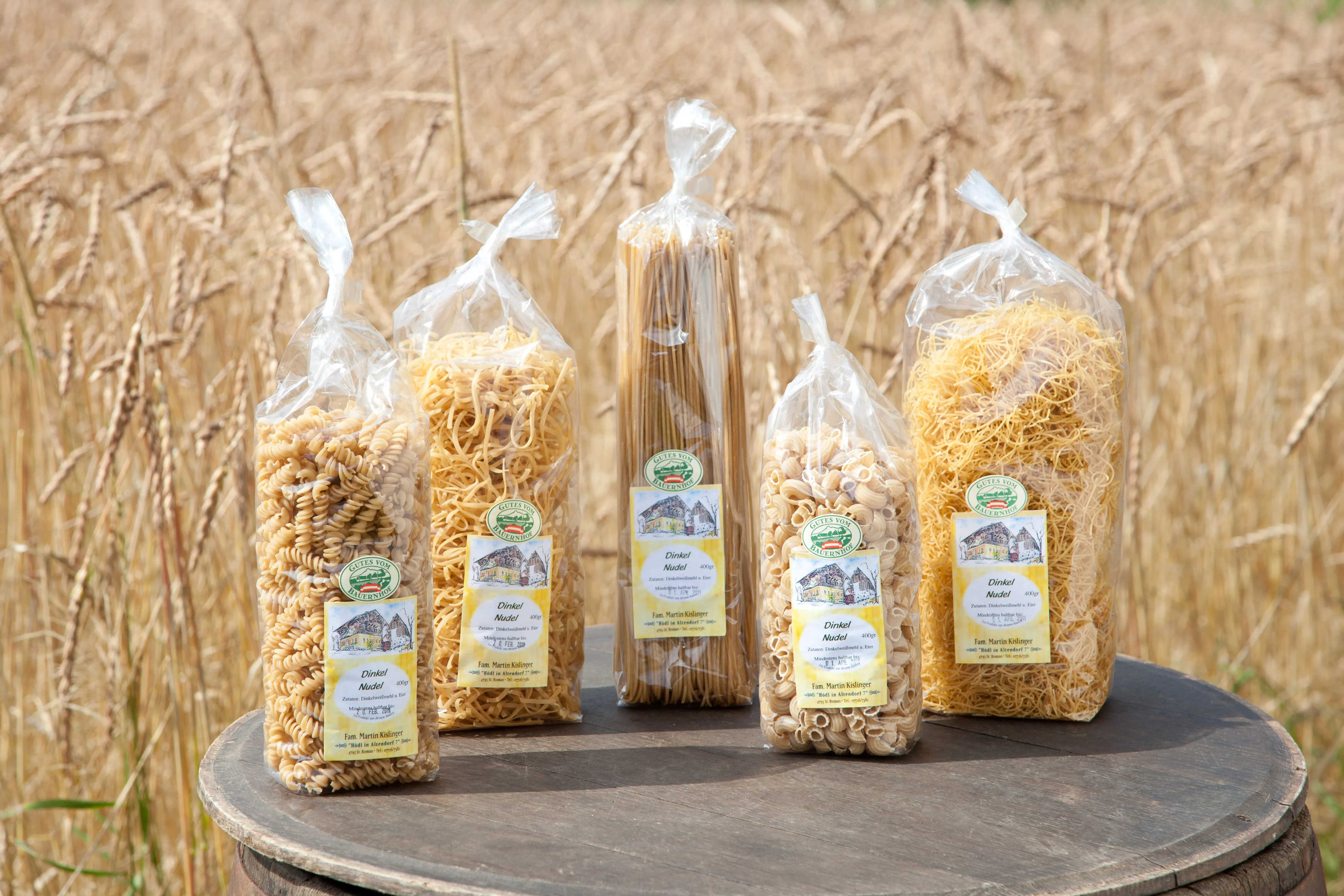 Auf einem Holzfass stehen fünf verschieden große Nudelpackungen mit verschiedenen Nudeln, dahinter Weizenfeld.