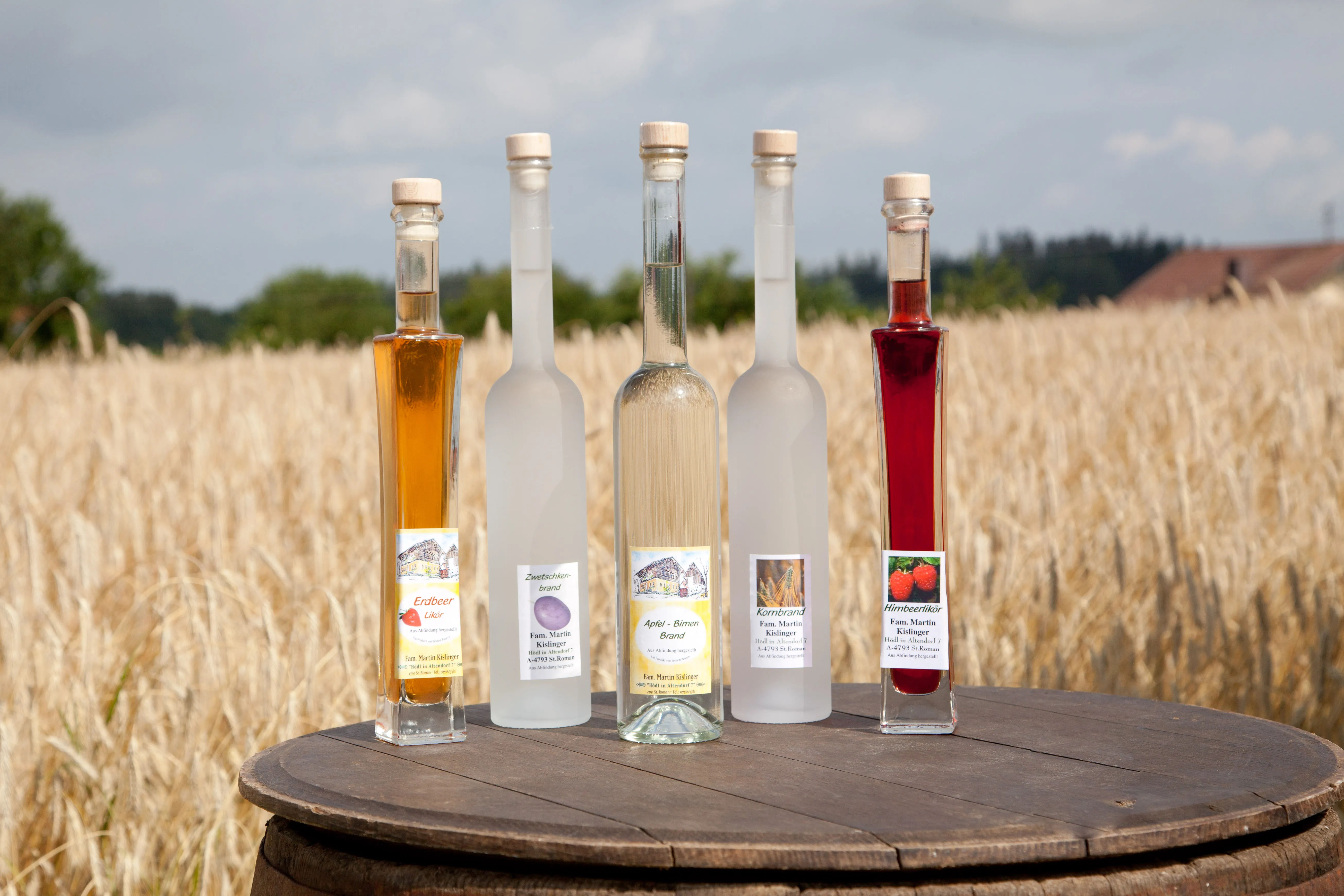 Auf einem Holzfass stehen fünf verschieden große Flaschen mit verschieden färbigen Spirituosen; dahinter ein Weizenfeld, grüner Wald und blauer Himmel