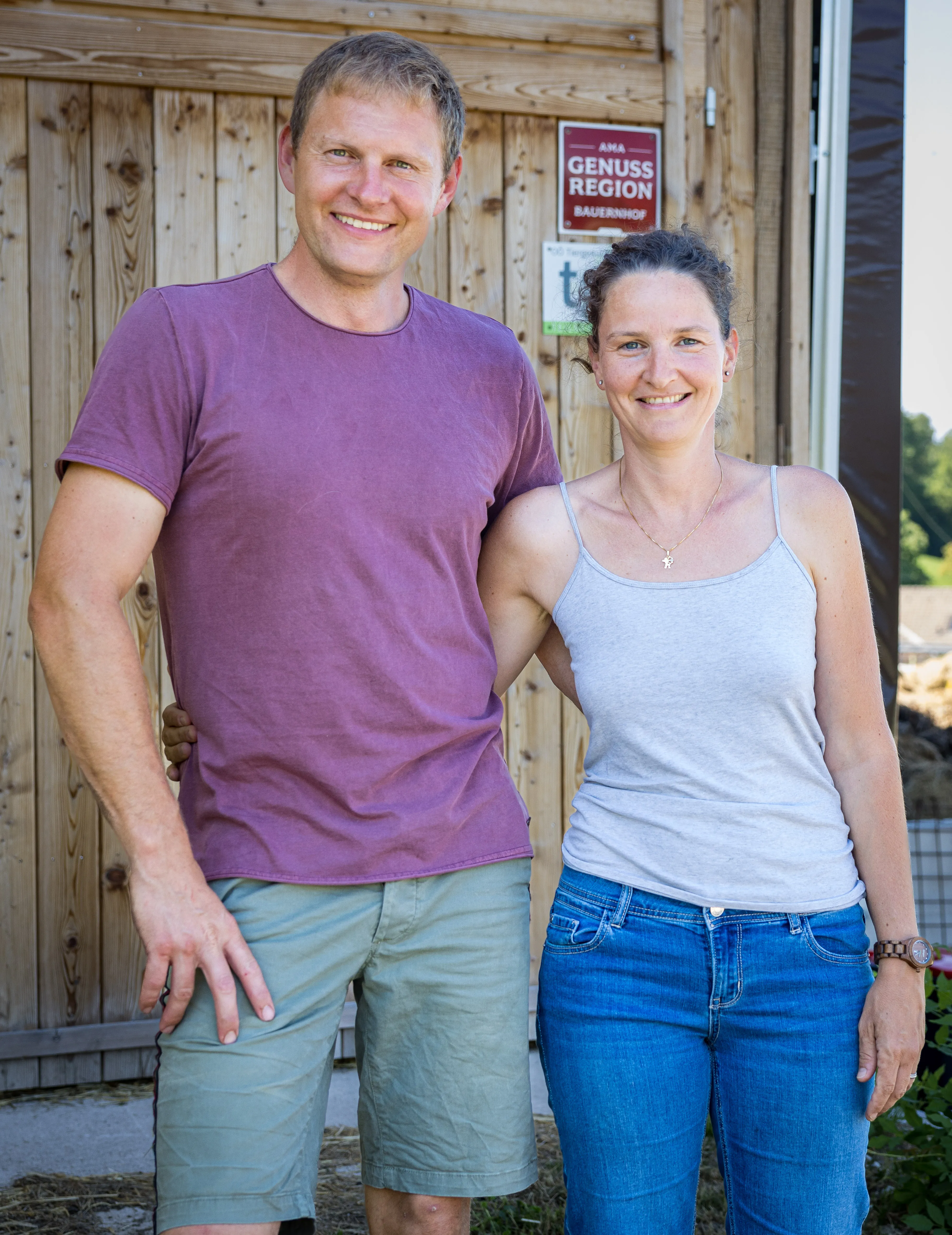 Eine Mann und eine Frau stehen vor einem hölzernen Hofladen, sie halten sich am Rücken umschlungen