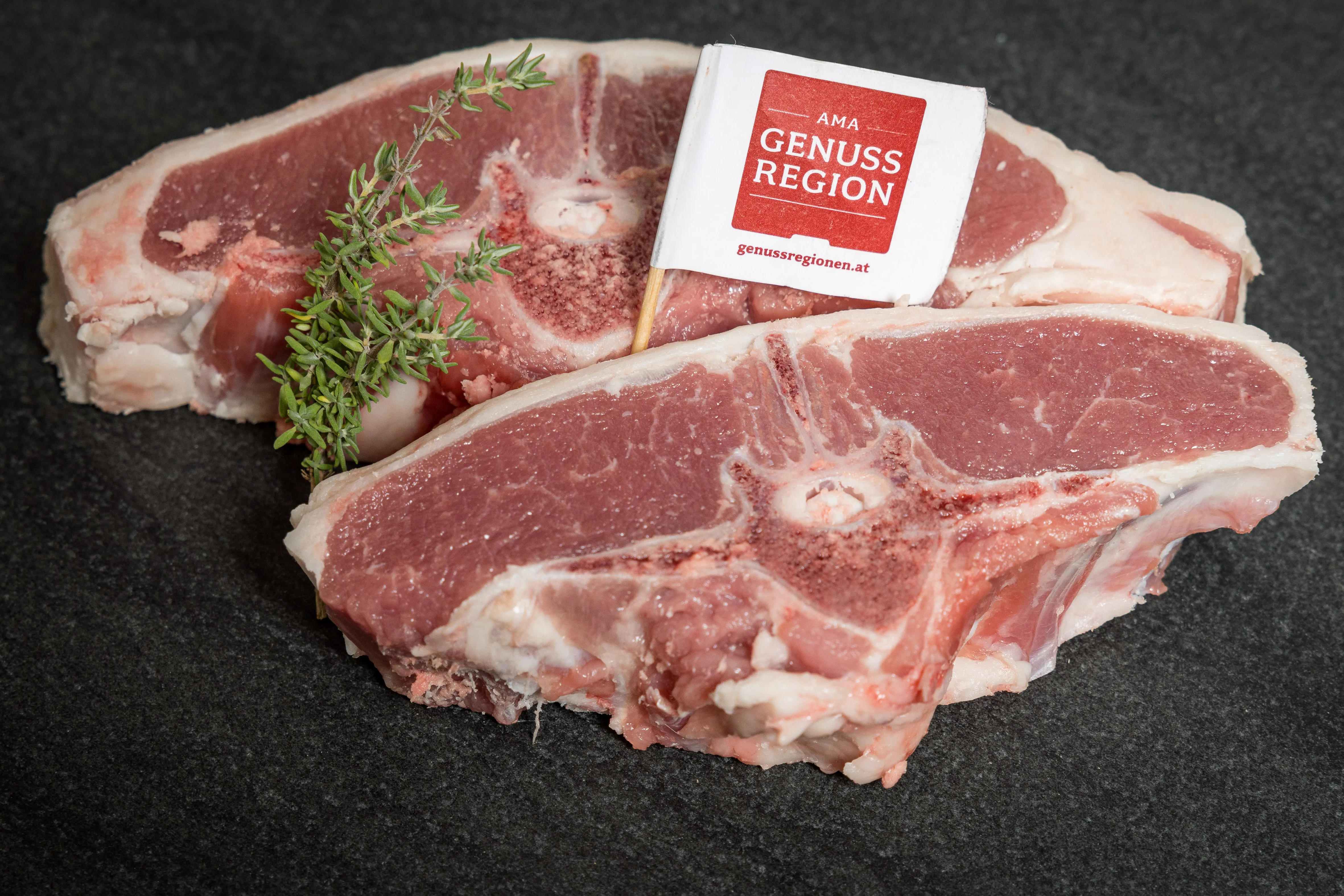 Auf einer schwarzen Platte liegen zwei Stück Frischfleisch, garniert mit Kräutern