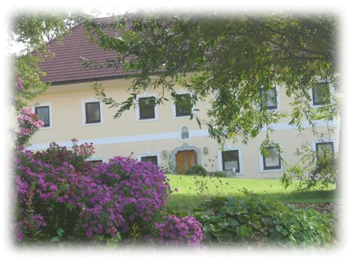 Man sieht ein gelbes Bauernhaus mit weißen Streifen und Fensterrahmen, davor grüne Bäume, lila Büsche und grüne Wiesen