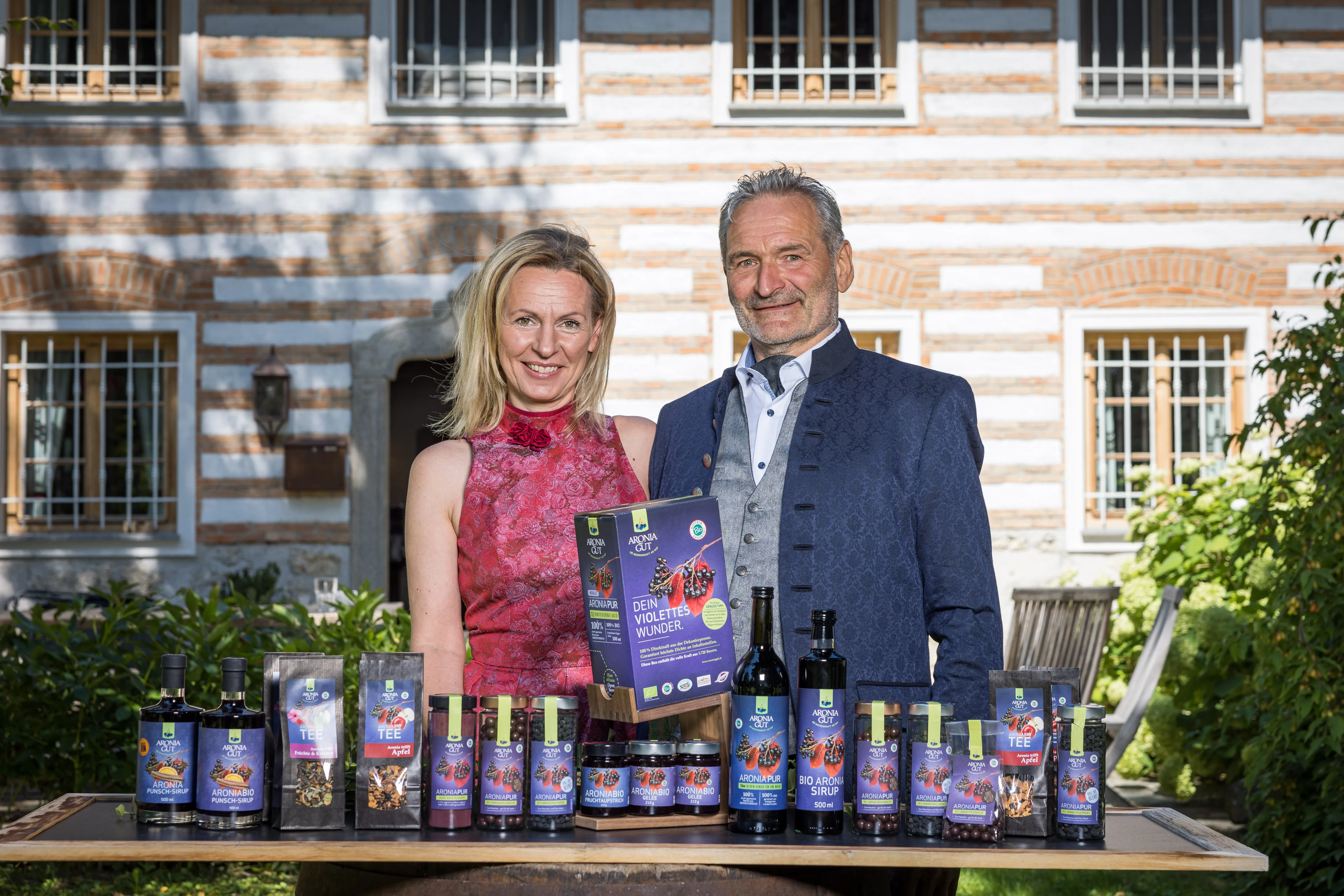 Herr und Frau Rogl stehen vor einem Bauernhaus; vor ihnen ein Tisch mit verschiedenen Aroniaprodukten in Form von Flaschen, Gläser und Sackerl.