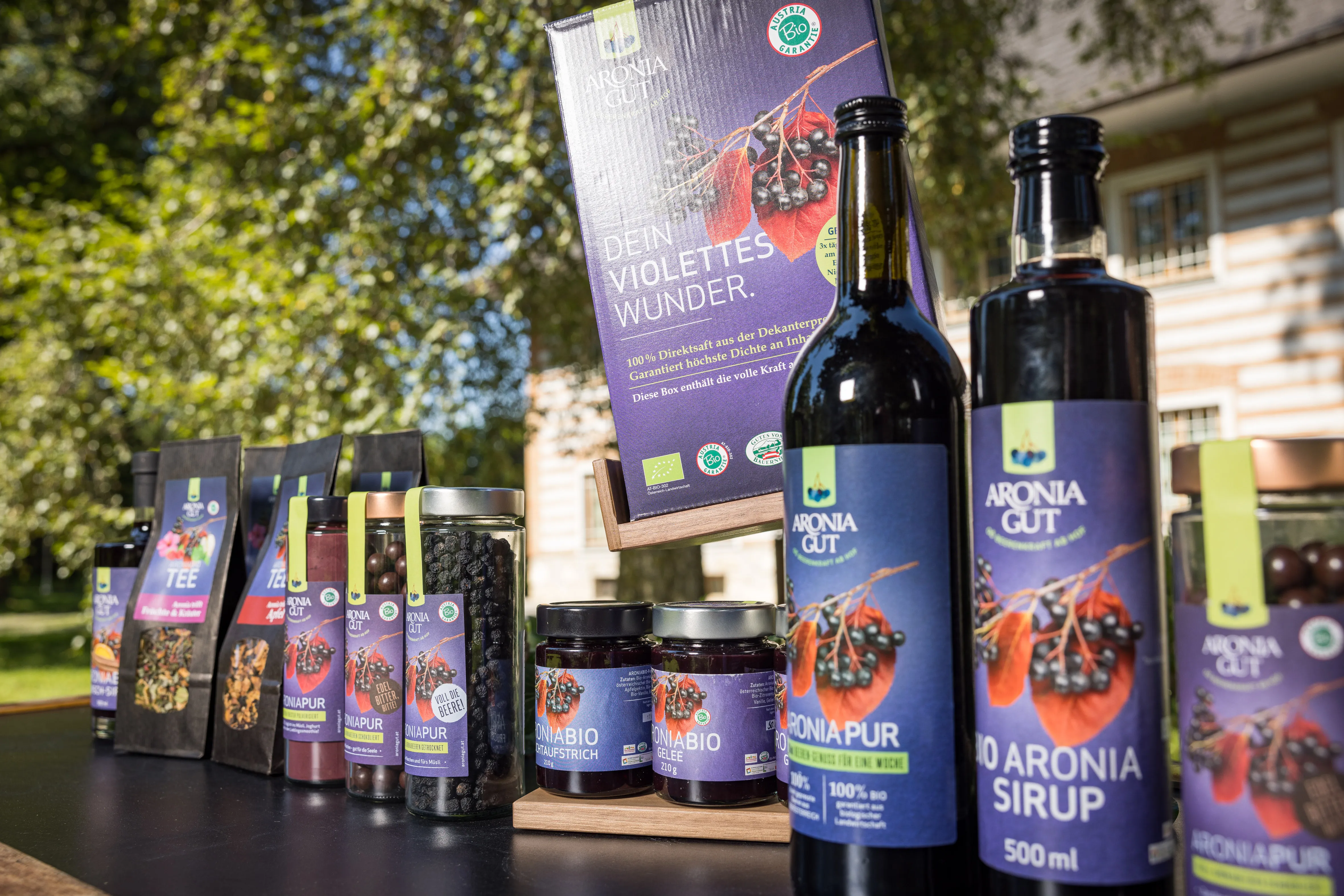 Auf dem Bild stehen mehrere Flaschen und Gläser mit lilafarbenen Etiketten mit Aroniasaft und ähnlichen Aroniaprodukten. Im Hintergrund Bäume.