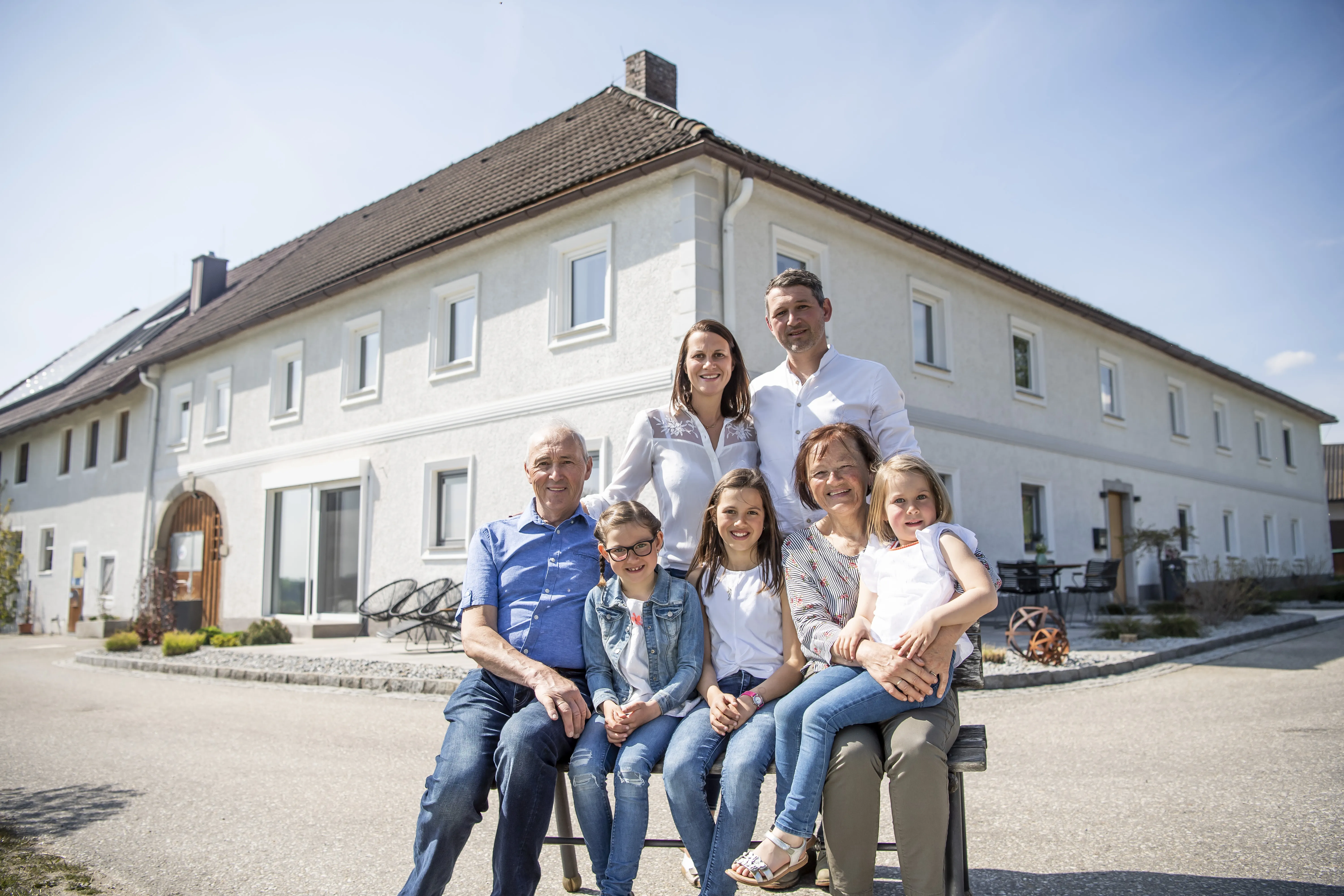 vor einem großen weißen Bauernhaus stehen bzw. sitzen ein älterer und ein jüngerer Mann, eine ältere und eine jüngere Frau und drei Mädchen. vor dem Haus ist der Vorplatz asphaltiert. Alle tragen weiße bzw. blaue Oberteile und Jeans. 