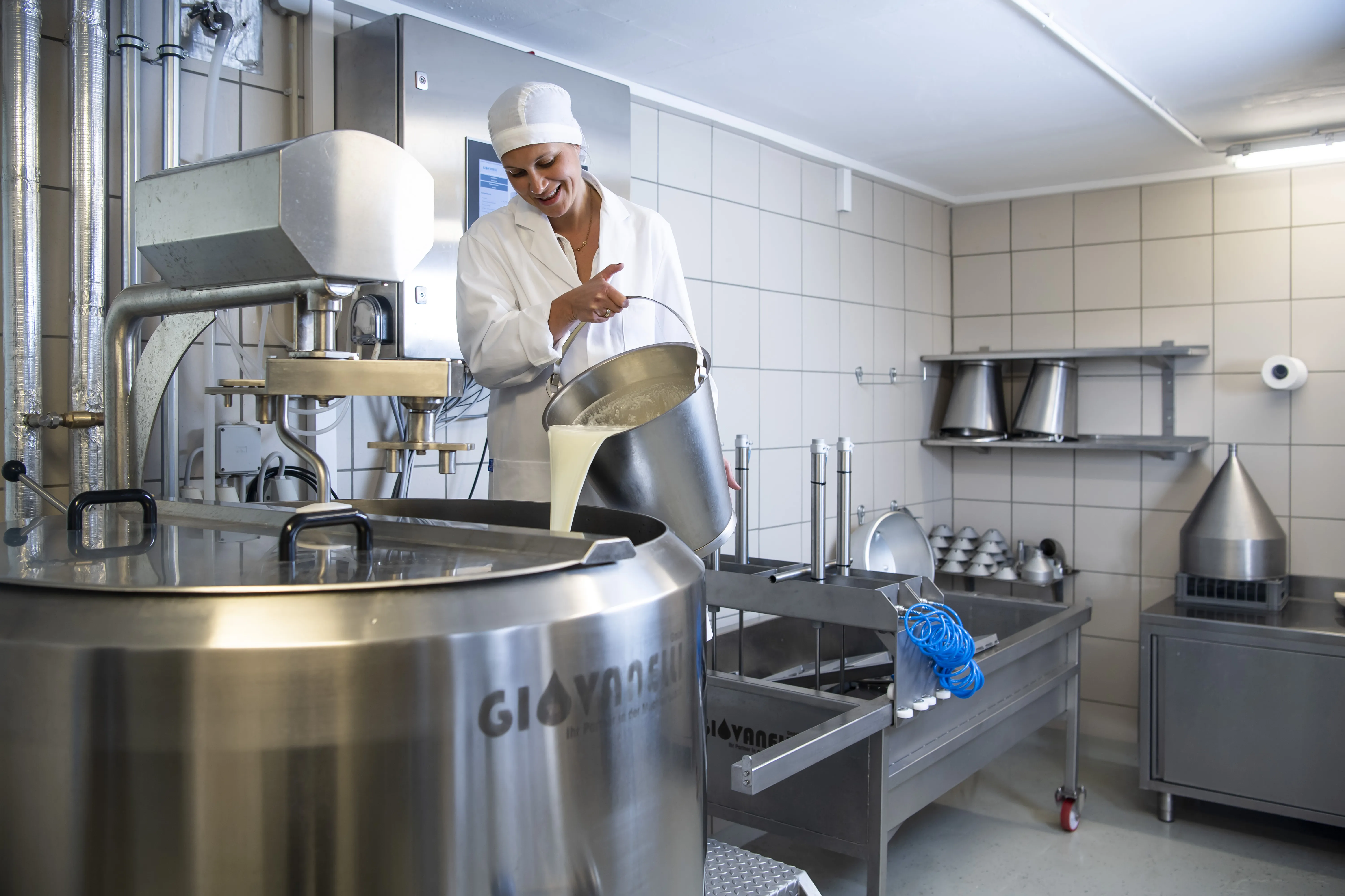 Auf dem Bild sieht man einen Verarbeitungsraum für Milch mit allen technischen Gerätschaften, daneben steht eine Frau in weißer Schutzkleidung und leer aus einem Blecheimer Milch in den großen Verarbeitungsbottich. Der Raum ist gefliest und hat eine weiße Decke.