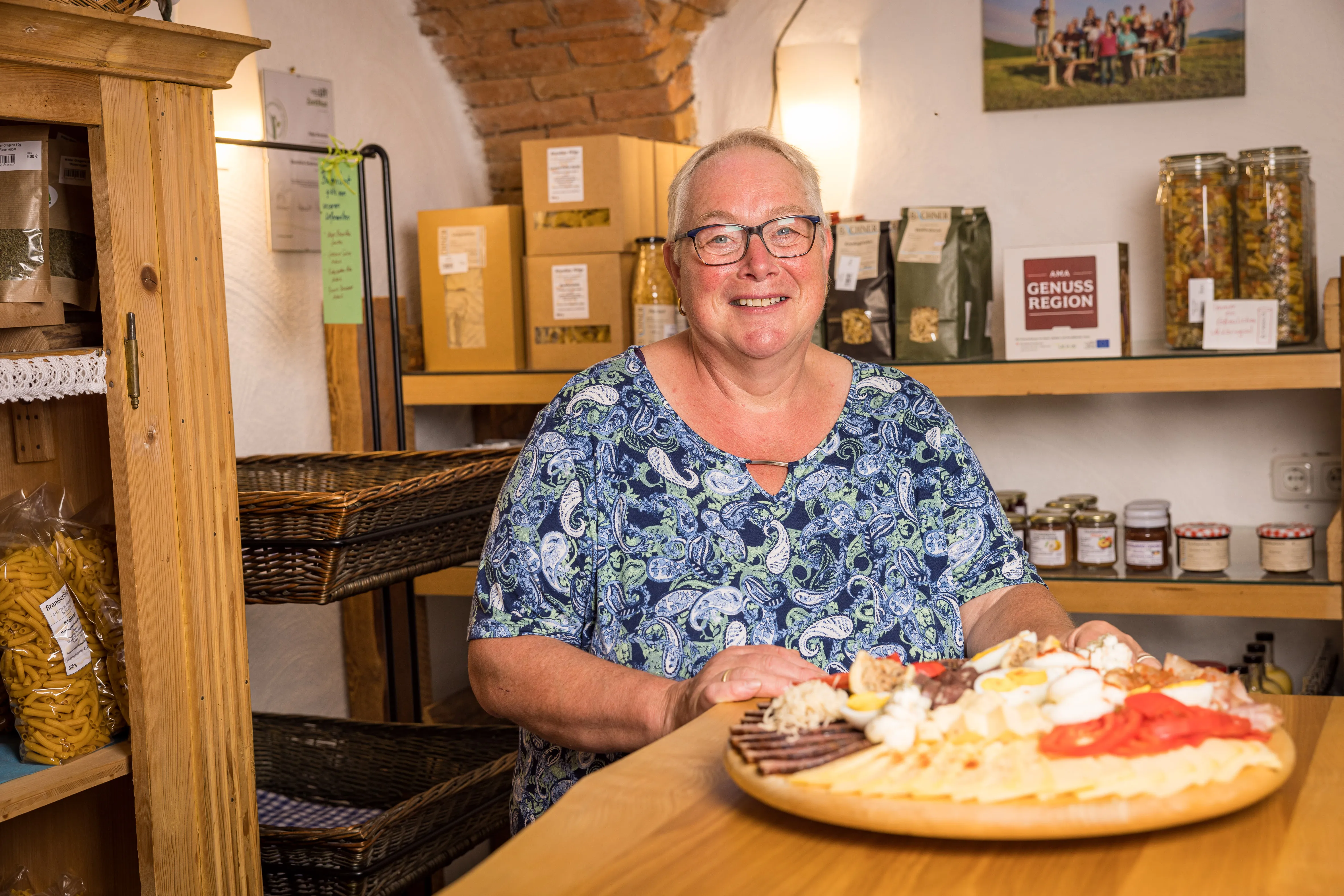 Frau Brandner steht im Hofladen. Vor ihr auf einem Tisch steht eine Platte mit Käse und Fleischprodukten. Dahinter Regale mit weiteren Produkten