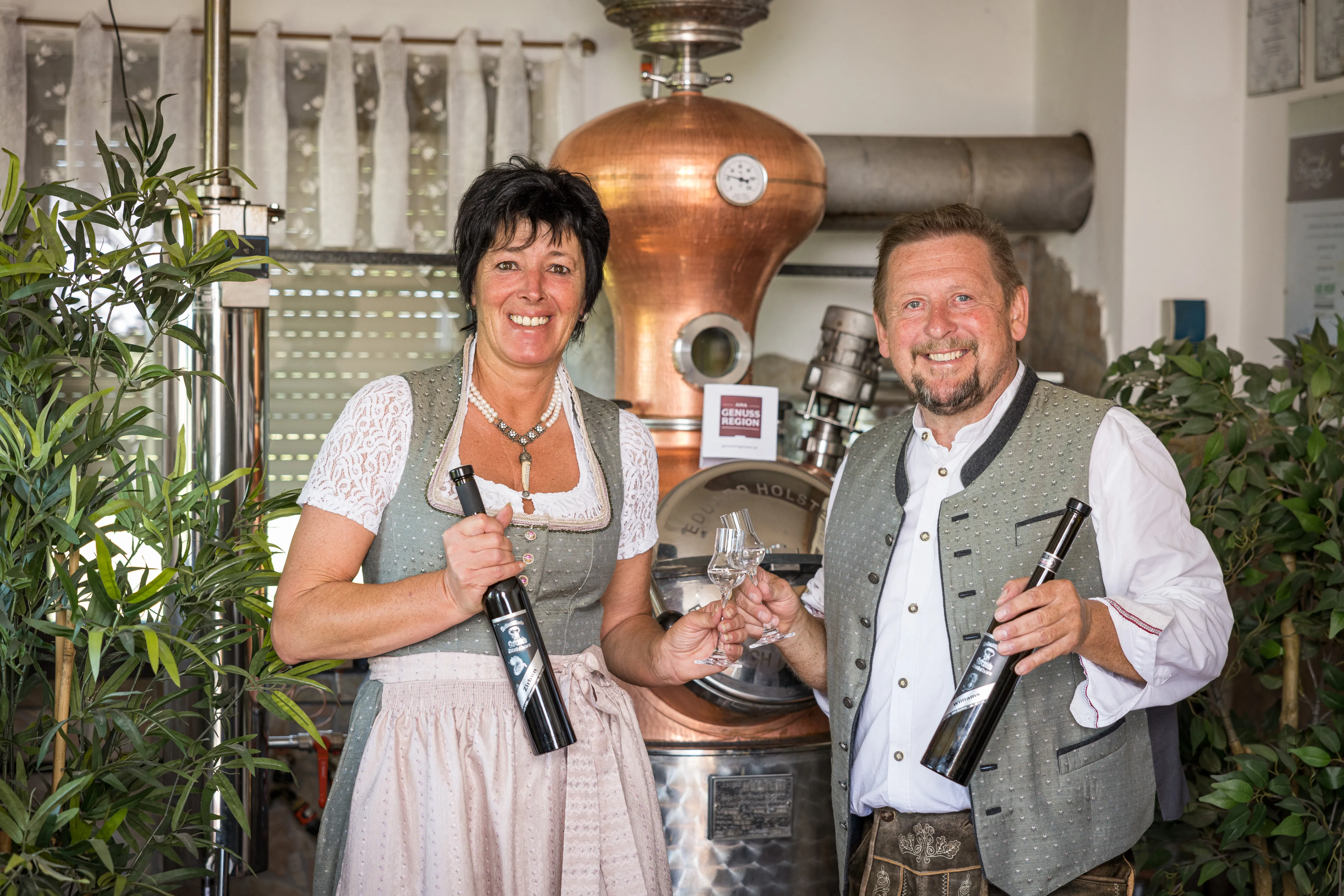 Herr und Frau Dicker, beide in Tracht, stehen vor einem Brennofen und halten jeweils ein Glas und eine Flasche in der Hand. Daneben Grünpflanzen