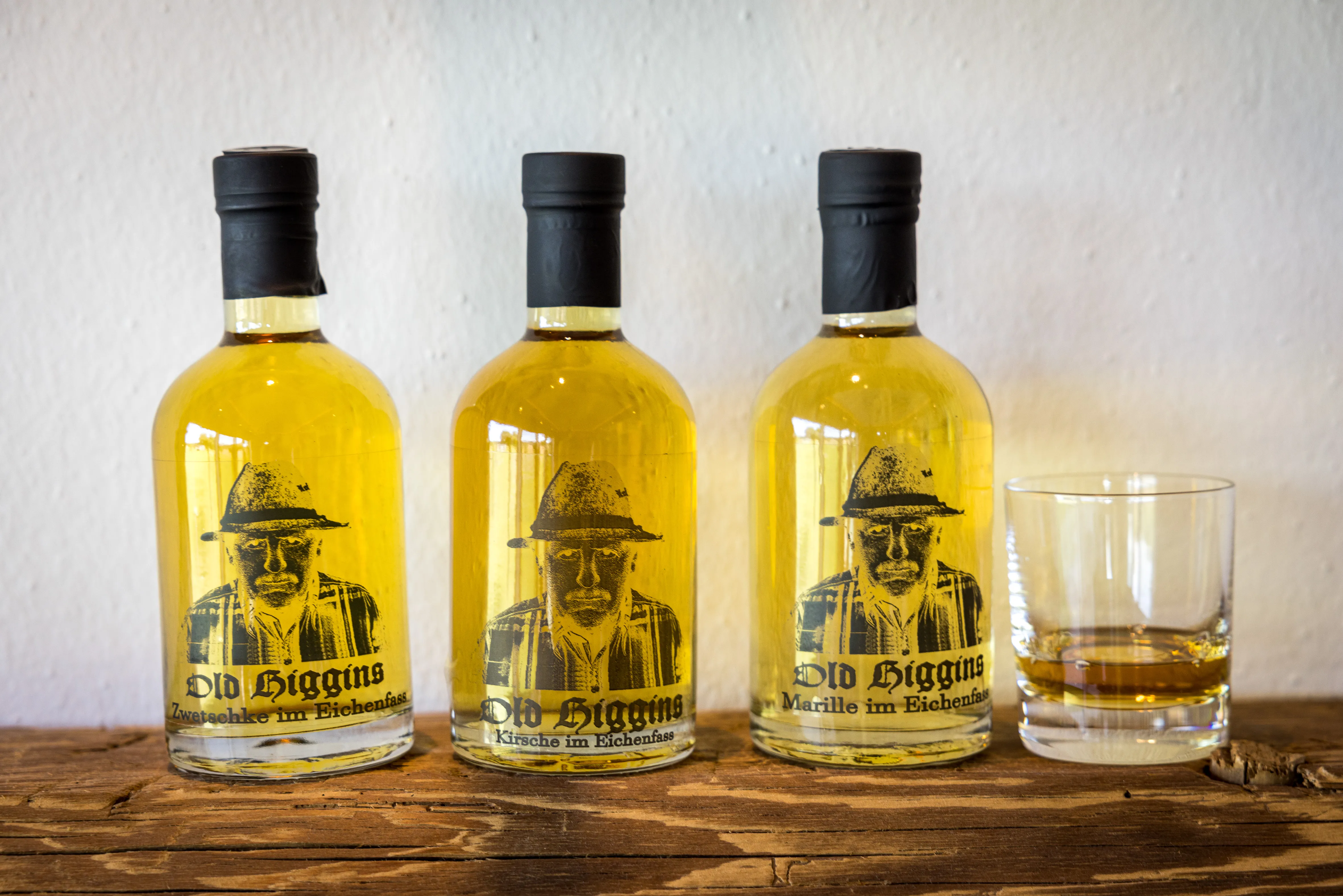Auf einer Holzplatte stehen drei Whiskyflaschen mit goldgelbem Inhalt und ein Glas mit Whisky, dahinter weiße Wand