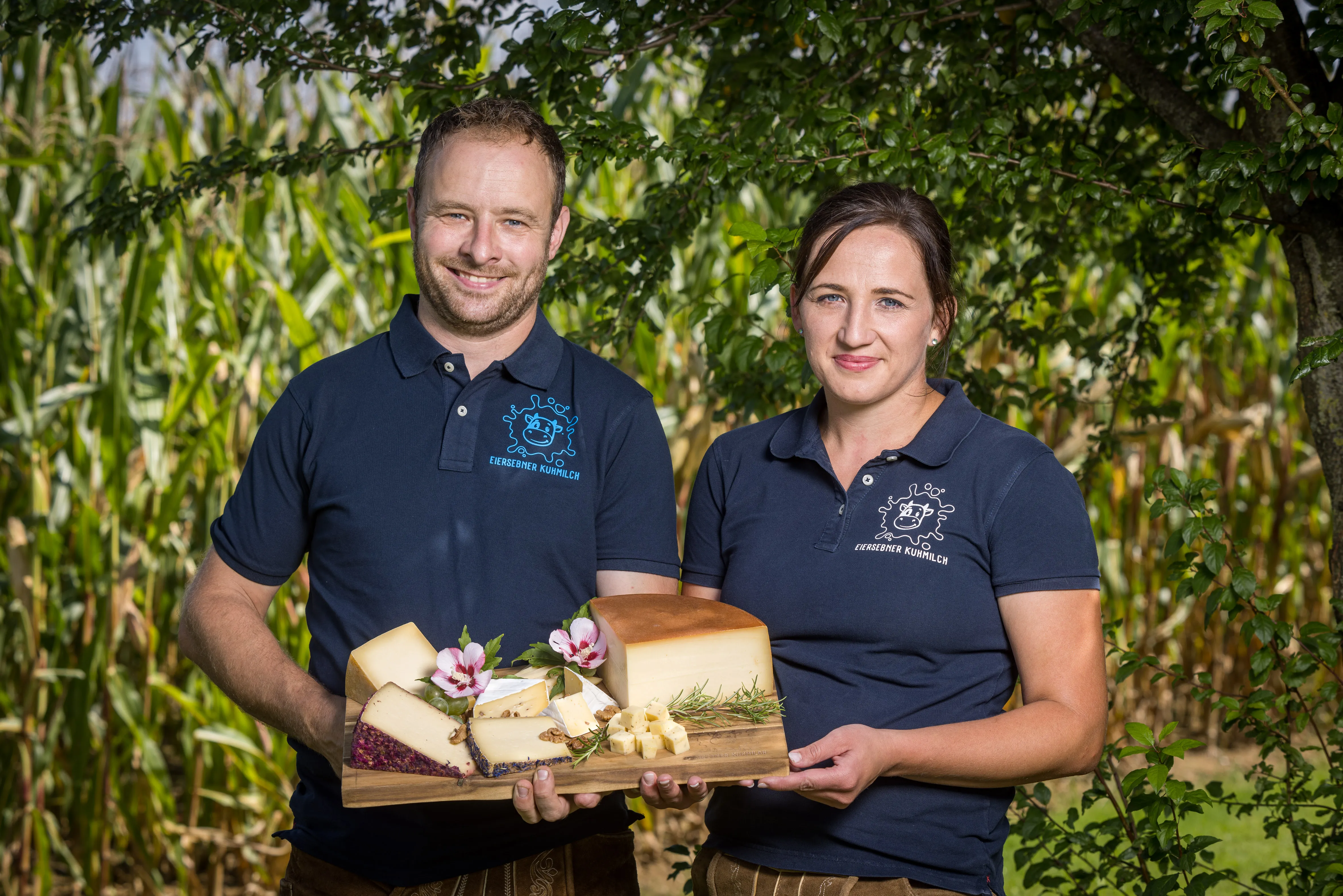 Ein Mann und eine Frau mit blauem Polo-Shirt halten gemeinsam ein großes Brett mit Käsespezialitäten in den Händen; dahinter grüne Bäume und Büsche
