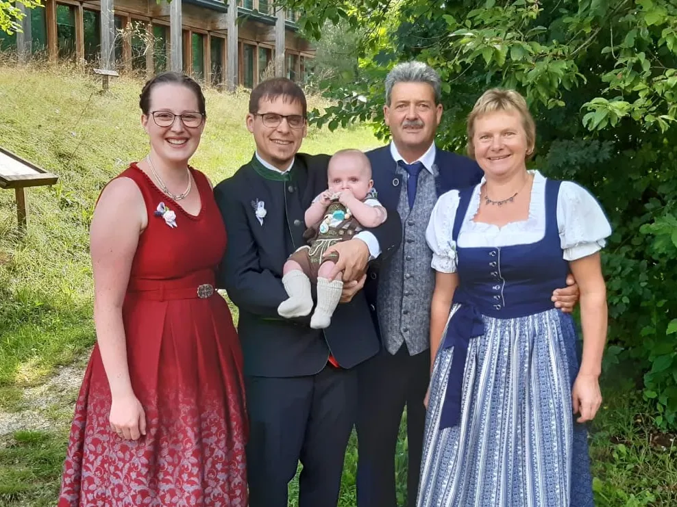 Zwei Frauen im roten und blauen Dirndl und zwei Männer im Anzug, wobei einer ein Kleinkind auf dem Arm hält, stehen auf einer Wiese, im Hintergrund ein Gebäude und Sträucher