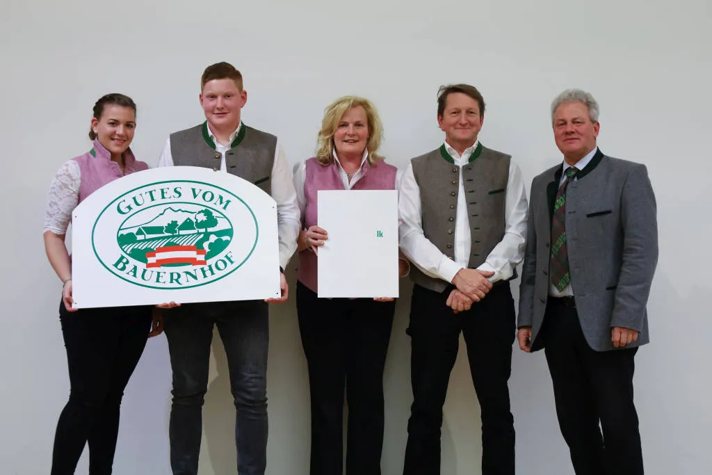 Familie Fischer, zwei Erwachsene, eine Tochter und ein Sohn stehen mit einem weiteren Mann vor einem weißen Hintergrund und halten eine Hoftafel und eine Urkunde in die Kamera