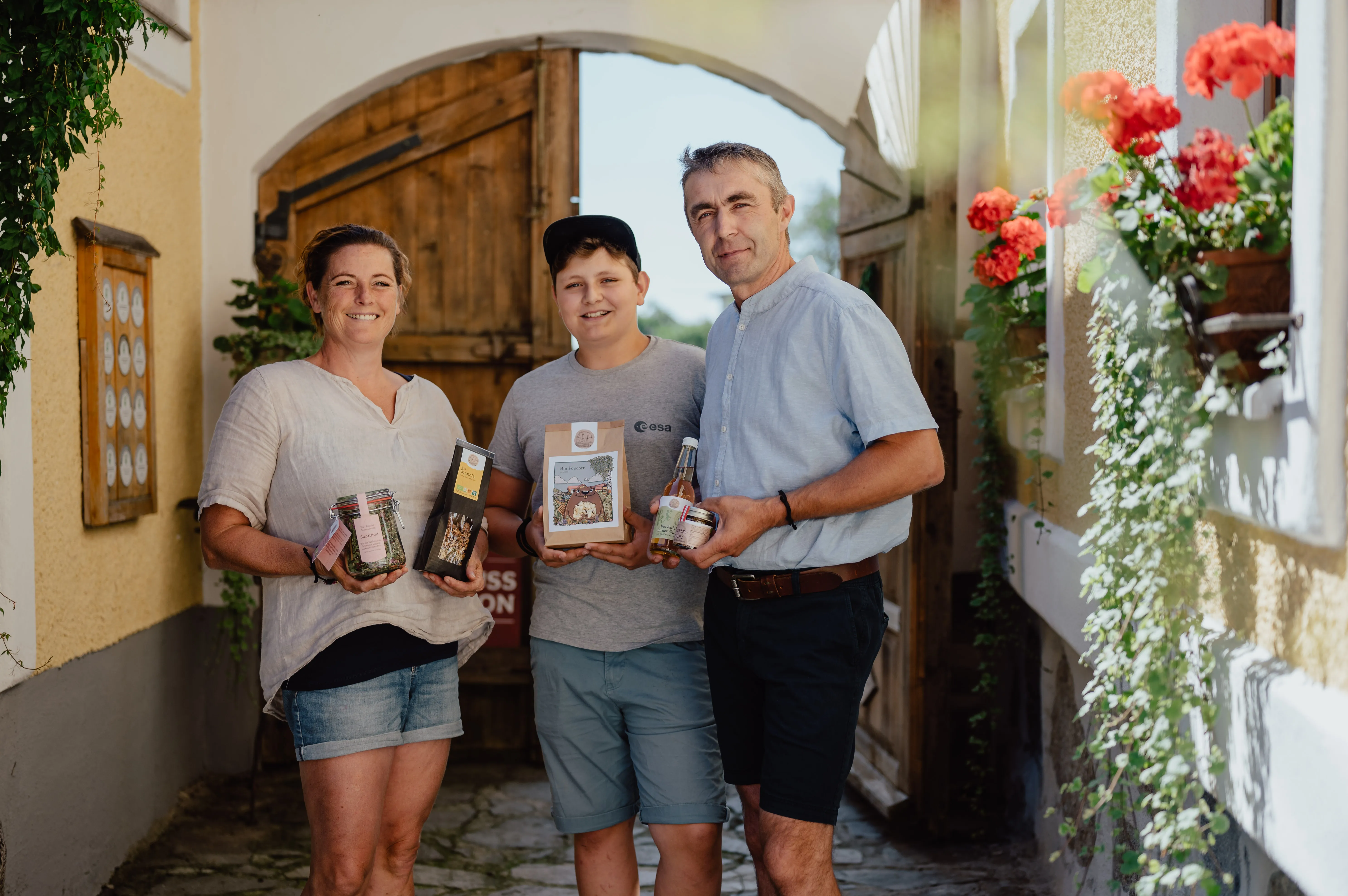 In einem Hofeingang stehen ein Mann, eine Frau und ihr Sohn. Sie halten verschiedene Produkte in den Händen. Daneben Blumen an den Fenstern.