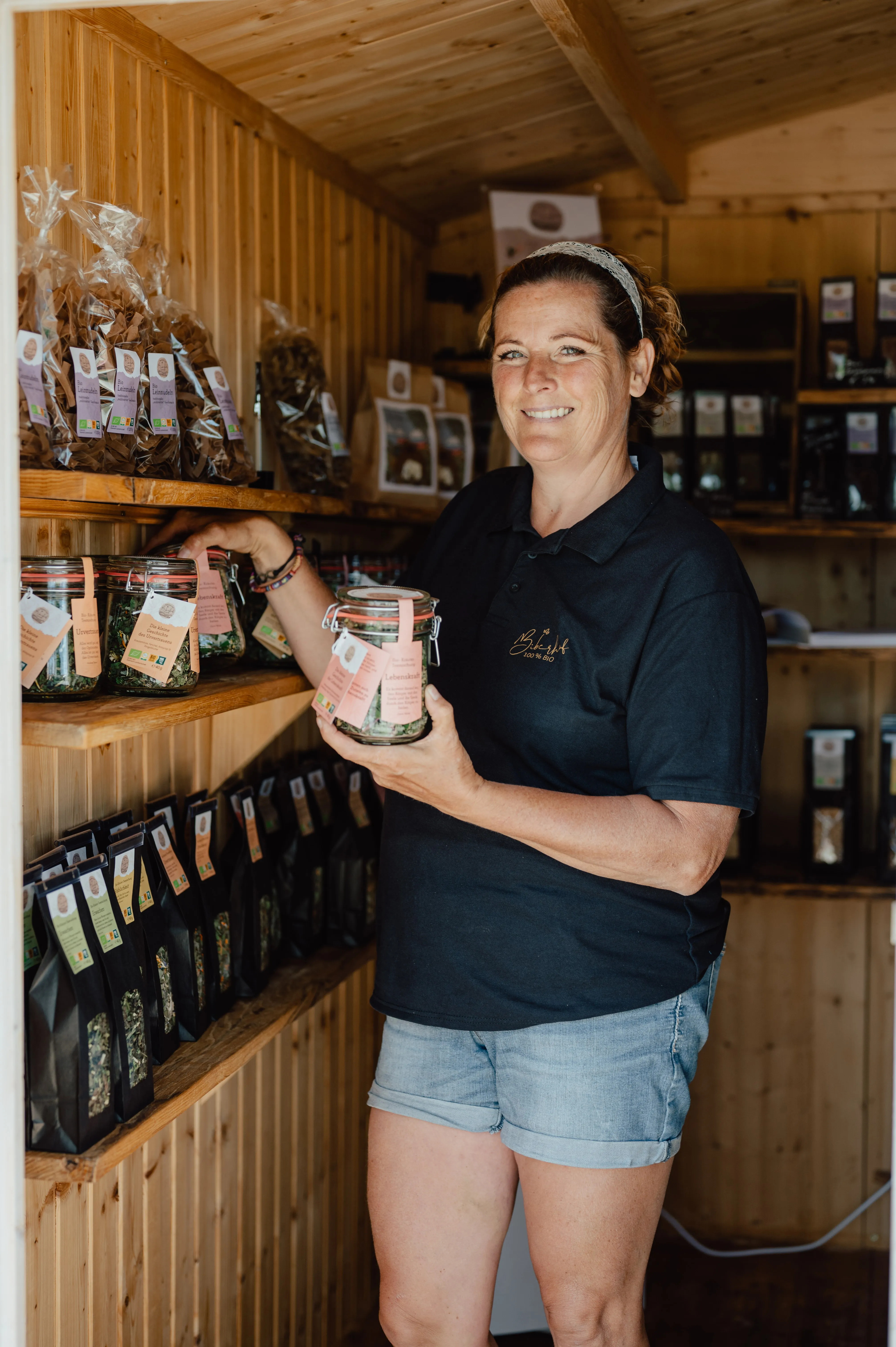 Eine Frau in dunklem T-Shirt und kurzer Hose steht in einem Hold-Verkaufsladen. An der Wand Regale mit verschiedenen Flaschen und Produkten