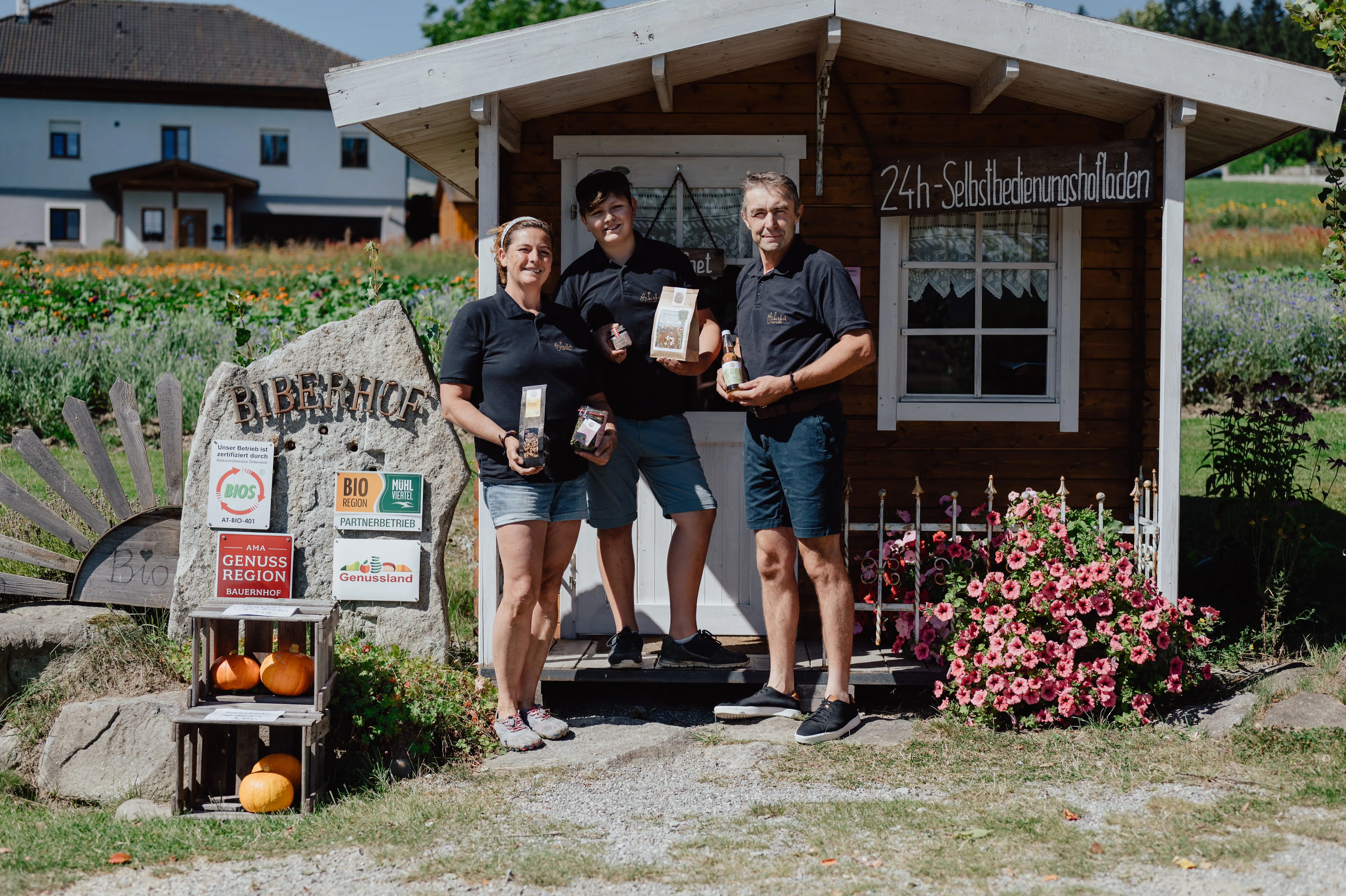 Vor einem Holzverkaufsstand stehen ein Mann, eine Frau und ein Junge und halten Produkte in den Händen. Daneben Kürbisse, Blumen und im Hintergrund ein Feld und ein Bauernhof