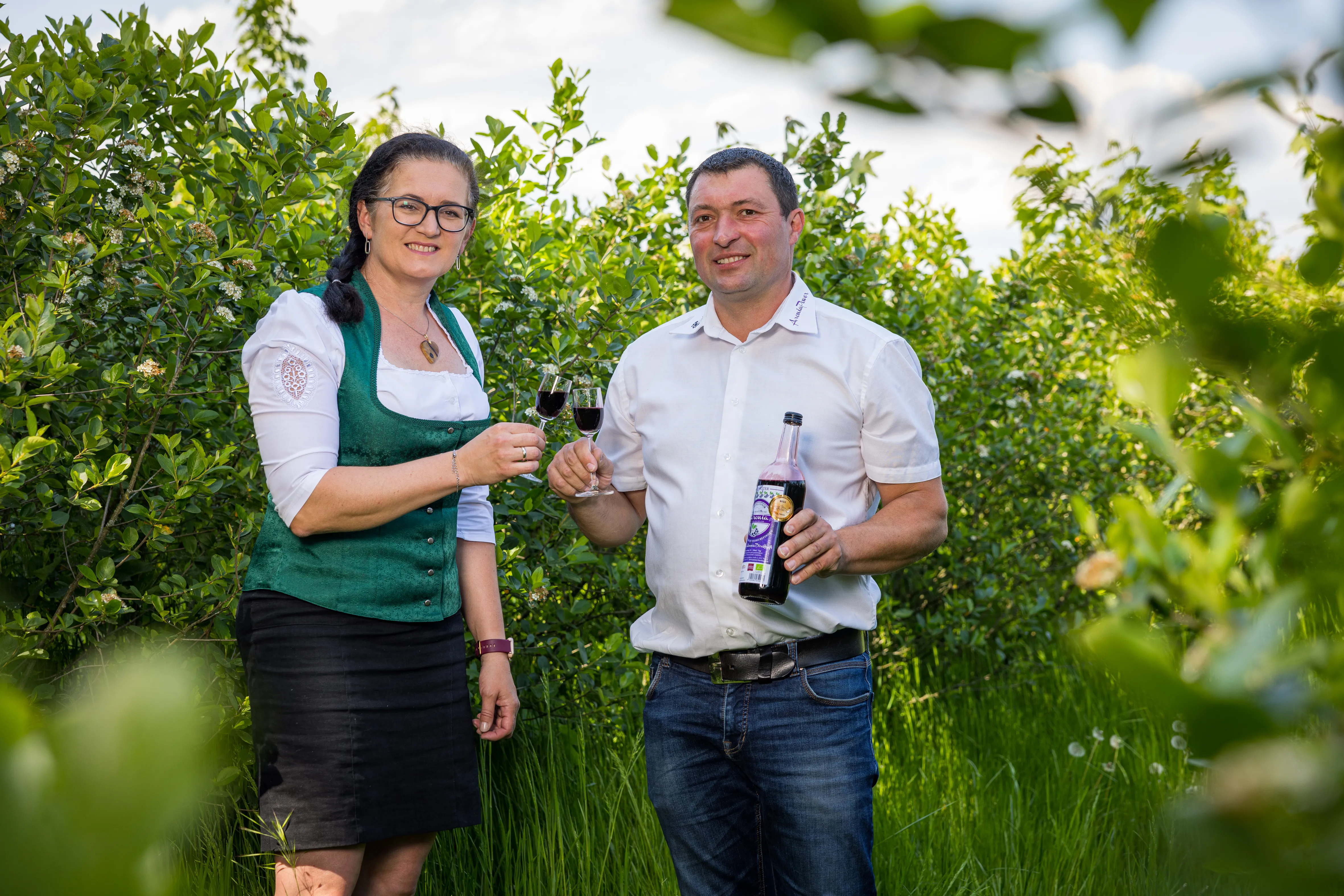 Ein Man und eine Frau stehen in einem Obstgarten und stoßen mit einem Aronialikör an. Herr Froschauer hält zudem eine Flasche in der Hand.