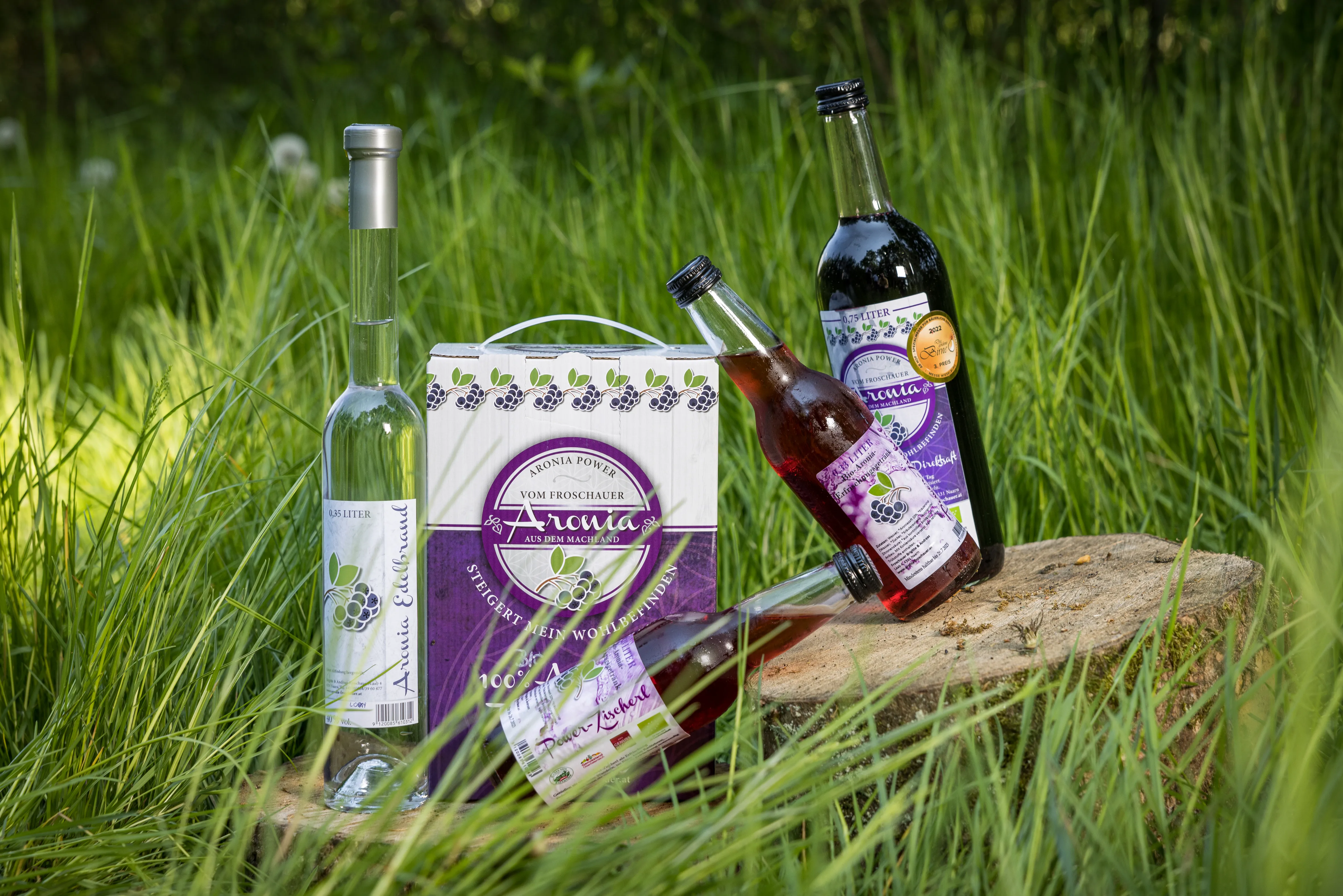 In einer grünen Wiese mit hohem Gras steht ein Holzbrett mit drei Flaschen Aronialikör und Aroniabrand und einem Papiersackerl.