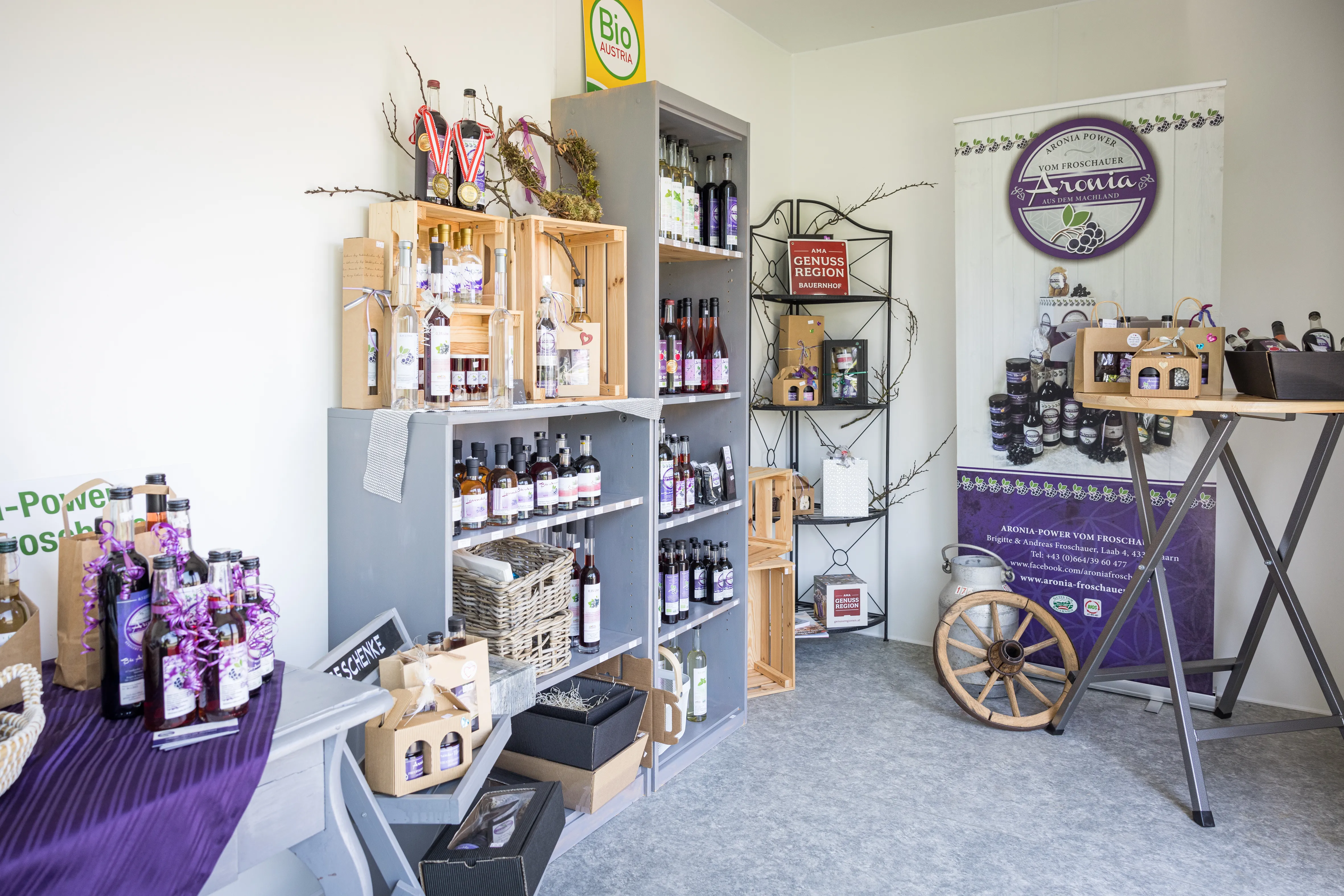 In einem weiß gestrichenen Raum sieht man graue und Holzregale mit Flaschen an der Wand stehen. Daneben ein Stehtisch und ein weiteres Eckregal. Dazwischen Dekormaterial.