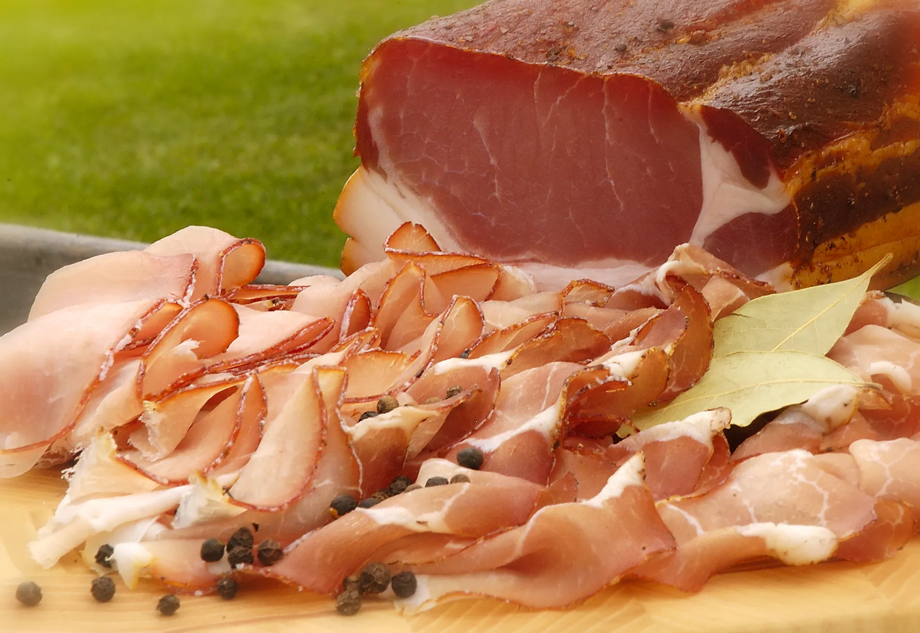 Auf einem Holzteller liegt frisch aufgeschnittener Speck und ein Stück Speck im Ganzen, dahinter grüne Wiese