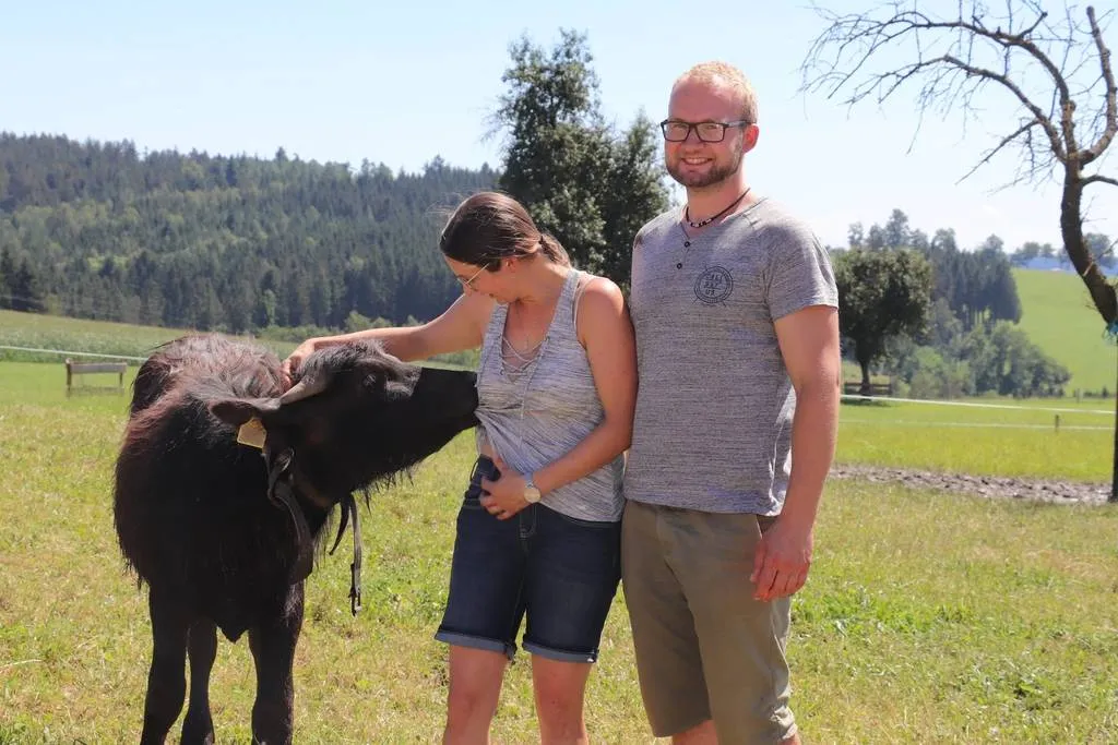 Ein Mann und eine Frau stehen auf einer Wiese; neben der Frau ein junger brauner Silberbüffel, der das Shirt der Frau im Maul hat. Dahinter grüne Bäume und Wiesen bzw. blauer Himmel