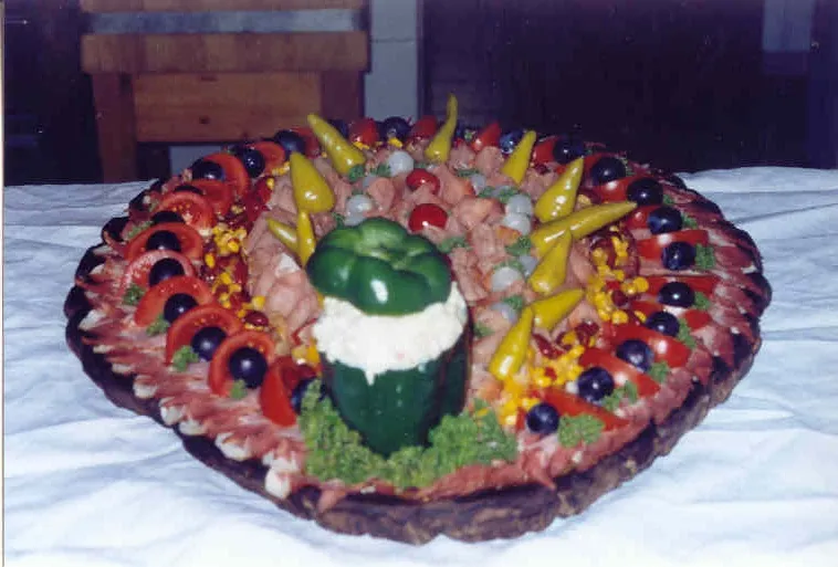 Auf einem Tisch mit weißer Tischdecke steht ein Jausenbrett mit Fleischprodukten, garniert mit Tomaten, Oliven, Paprika und Pfefferoni