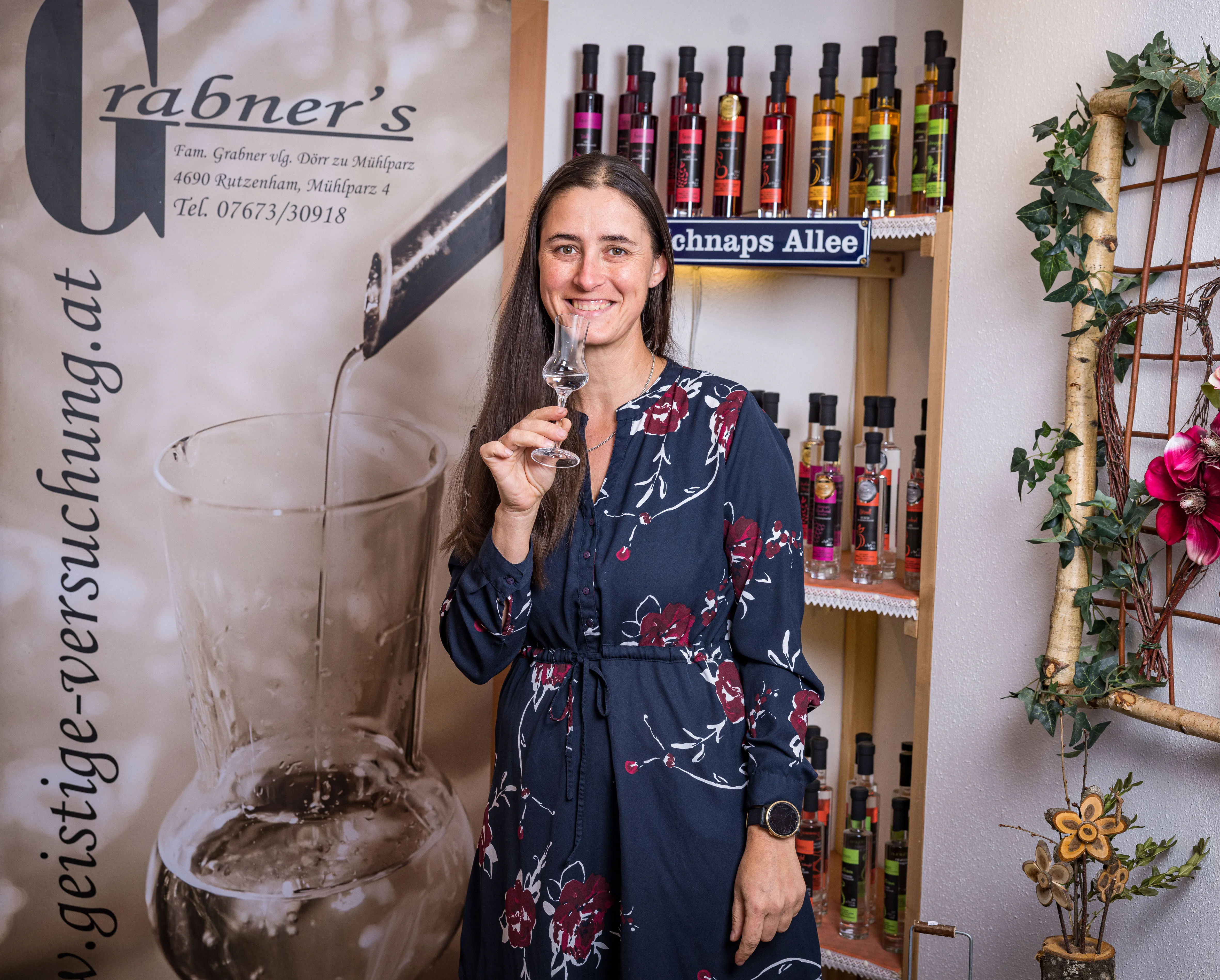 Eine Frau mit blauem Kleid hält ein Glas in der Hand und lächelt in die Kamera. Dahinter Holzegale mit verschiedenen Flaschen an der Wand.