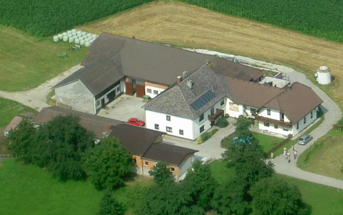 Aus der Vogelperspektive sieht man einen großen Bauernhof mit weißer Fassade, daneben Wirtschaftsgebäude, grüne Bäume und Sträucher und Wiesen, Felder und Heuballen