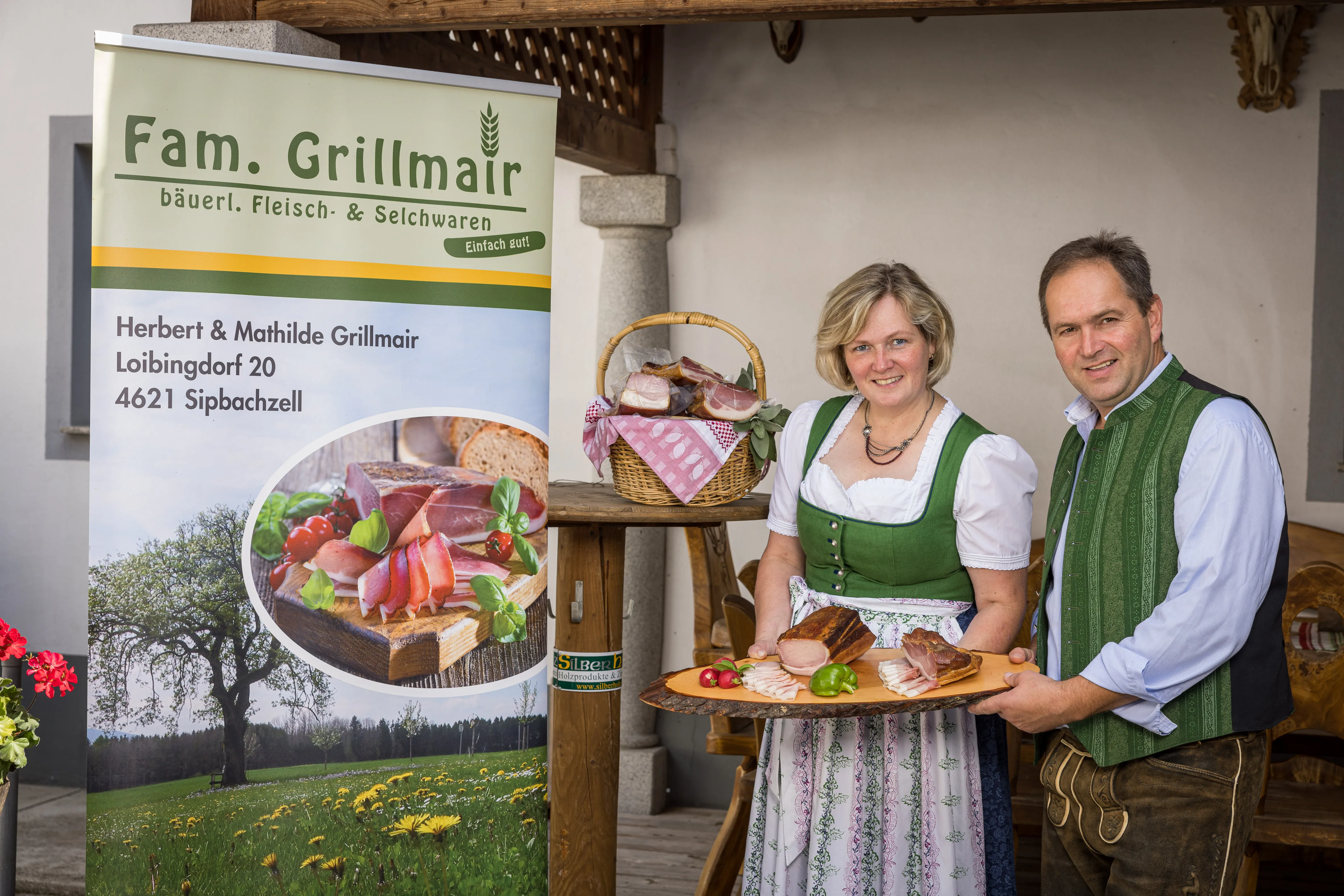 Ein Mann im Anzug und eine Frau im Dirndl halten eine große Platte mit verschiedenen Fleisch- bzw. Speckprodukten in den Händen. Daneben eine große Tafel mit ihrem Namen und Anschrift. Dahinter auf einem Stehtisch ein Korb mit weiteren Produkten