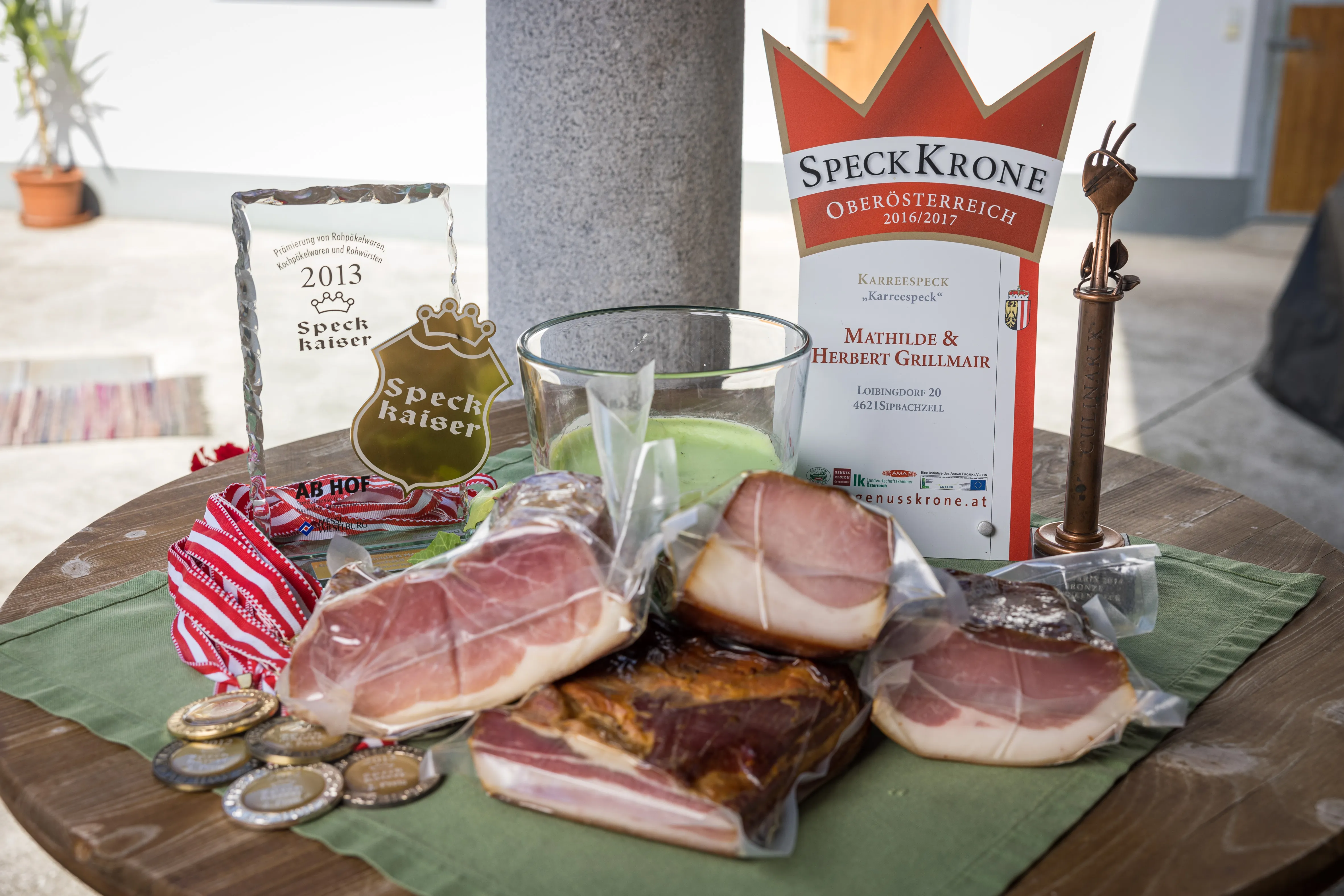 Auf einem Tisch liegen in Folie verschweißte Speckprodukte, daneben Medaillen und Auszeichnungen.