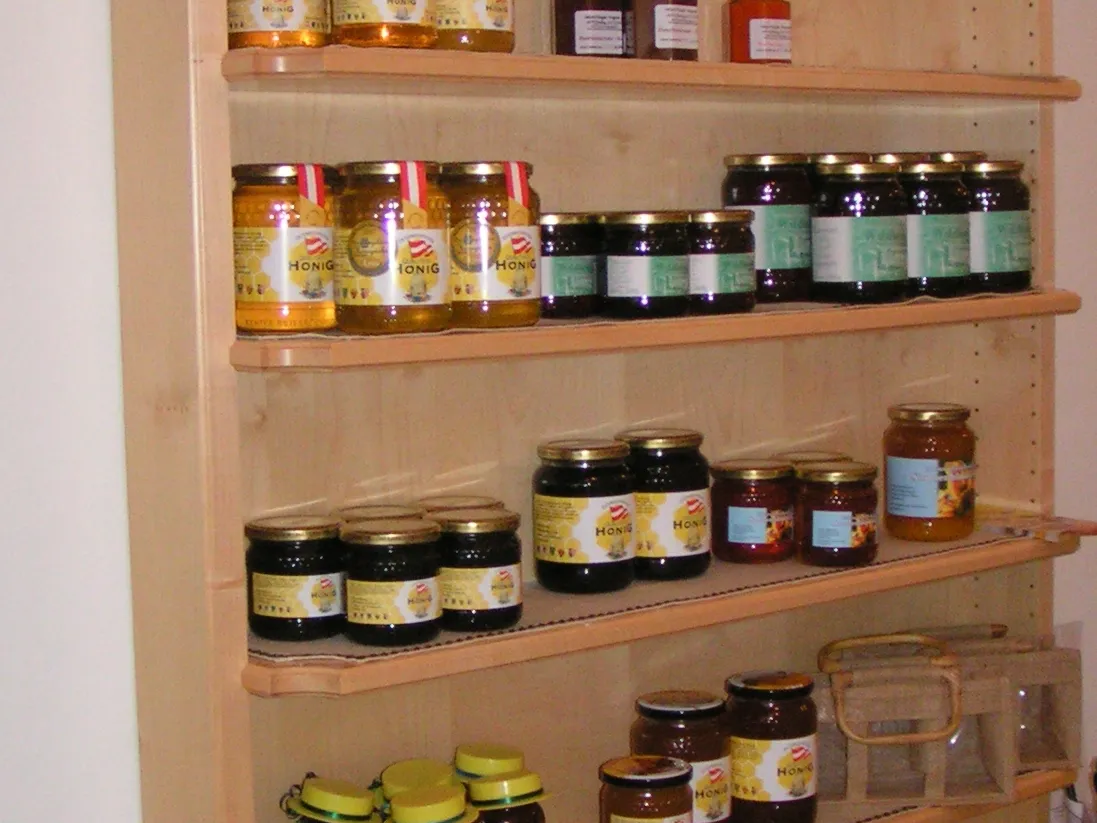 An einer weißen Wand steht ein Holzregal mit verschiedenen Honigprodukten, wie Gläser, Fläschchen...