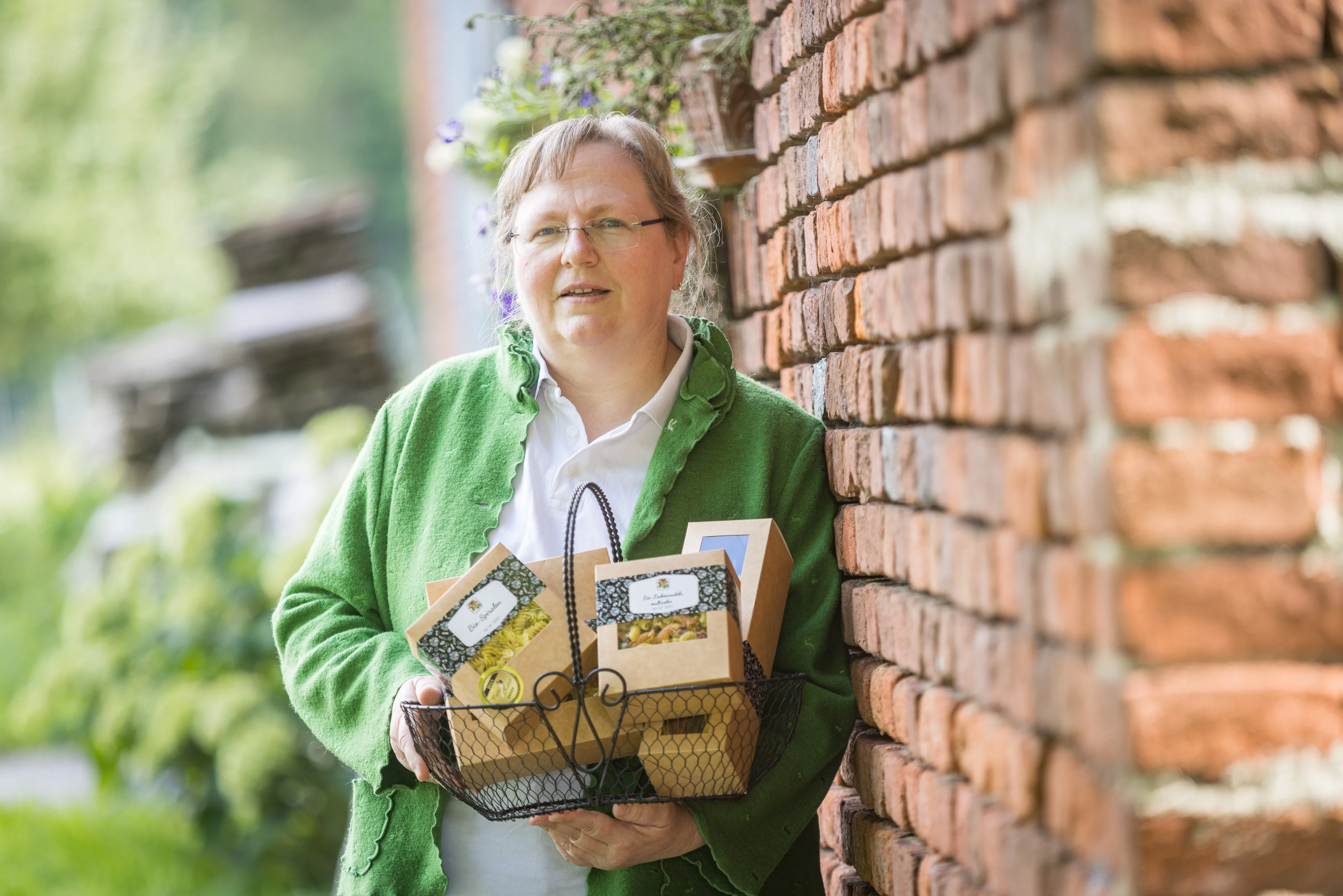Eine Frau mit grüner Weste und Brille lehnt an einer roten Steinmauer und hält einen Metallkorb mit verschiedenen Produktpackungen in den Händen. 