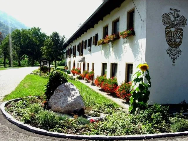 Auf dem Bild sieht man ein großes weißes Bauernhaus mit vielen Fenstern und Blumenkästen, mit wunderschönen Blumenschmuck, davor Wiese und ein Weg; daneben auch noch Bäume.