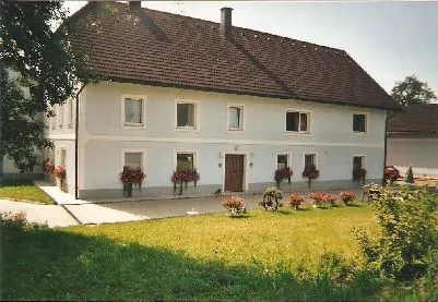 auf dem Bild sieht man ein großes helles Bauernhaus mit weißen Fenstern, rundherum Wiese und Wald