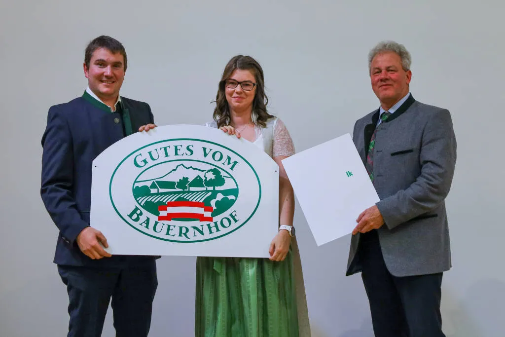 Herr und Frau Hölzl stehen mit einem weiteren Mann vor einem weißen Hintergrund und halten eine Hoftafel in den Händen. Der weitere Mann hält die Urkunde.